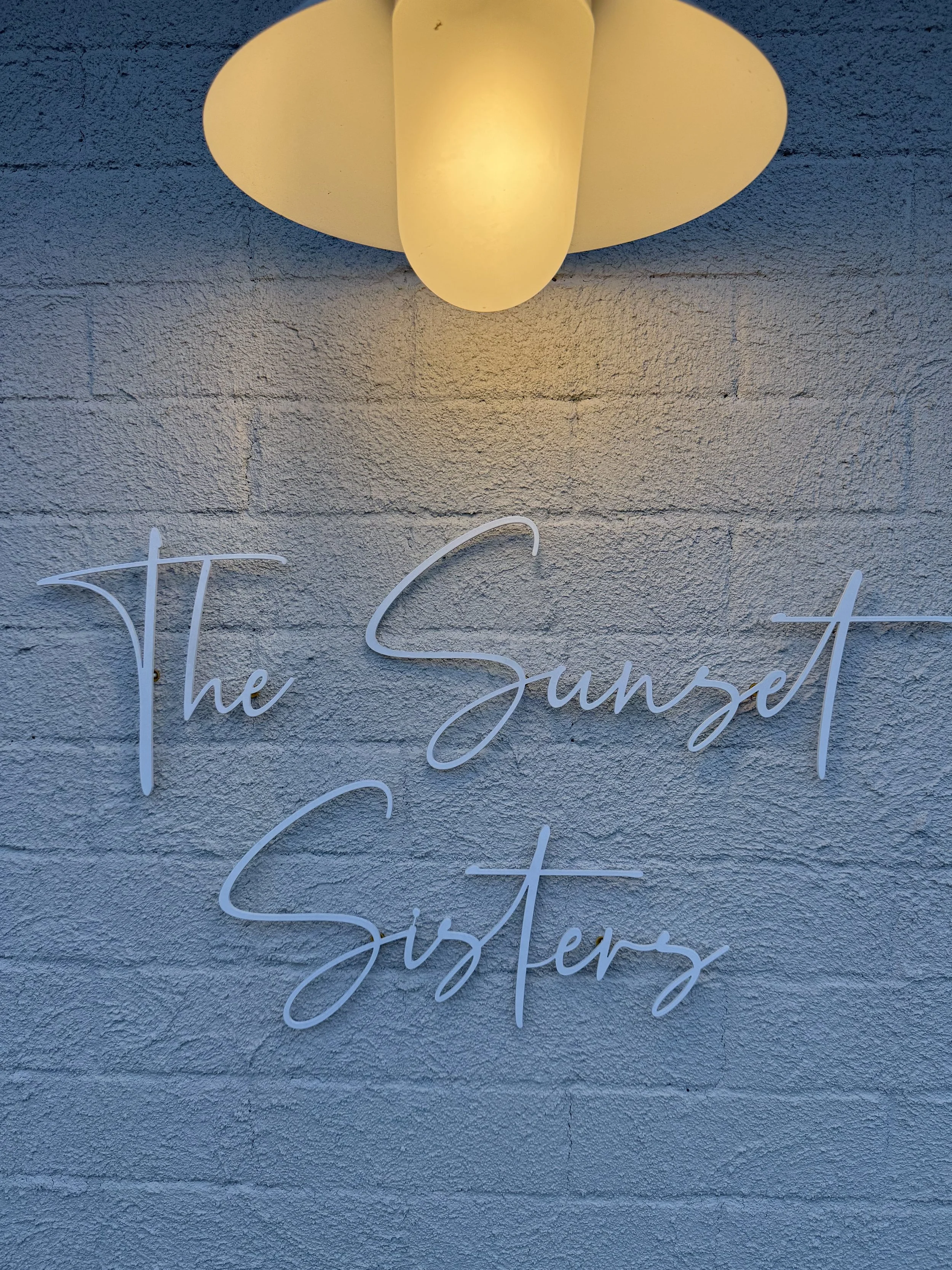 Neon sign reading 'The Sunset Sisters' mounted on a textured concrete wall, illuminated by a ceiling light fixture above.