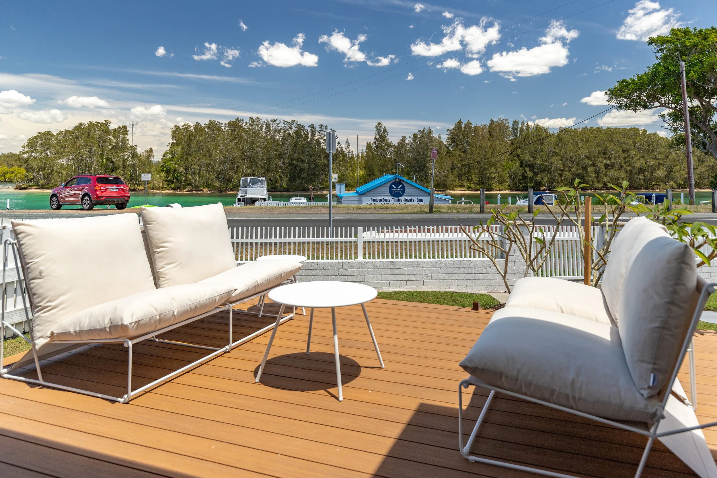 Outdoor patio with two white cushioned chairs and a white round table on a wooden deck, overlooking a canal with boats, trees, and a blue boat rental shop in the background.