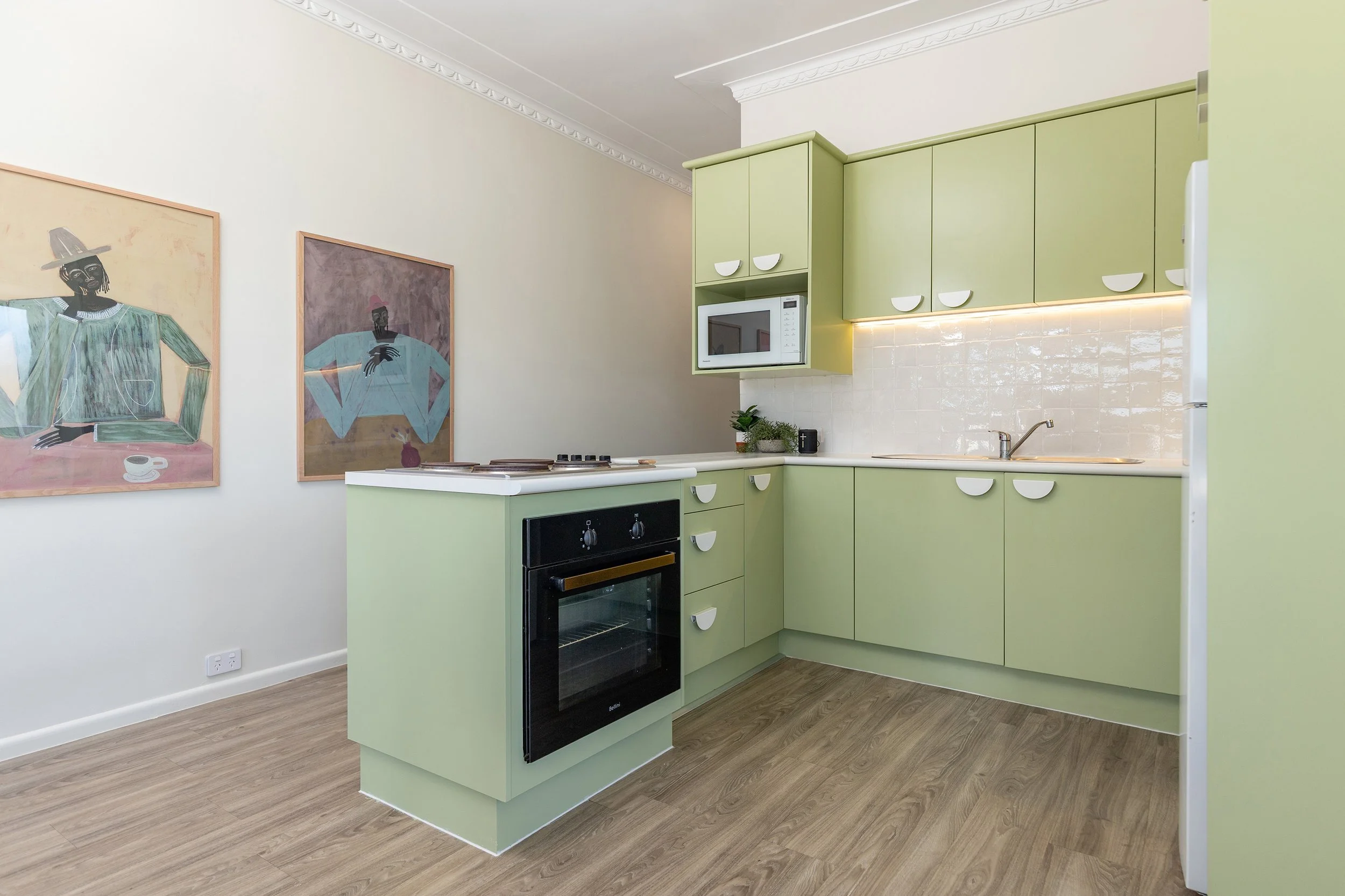 Kitchen with light green cabinets, wooden flooring, a black oven, microwave, white tiled backsplash, plants, and wall art.