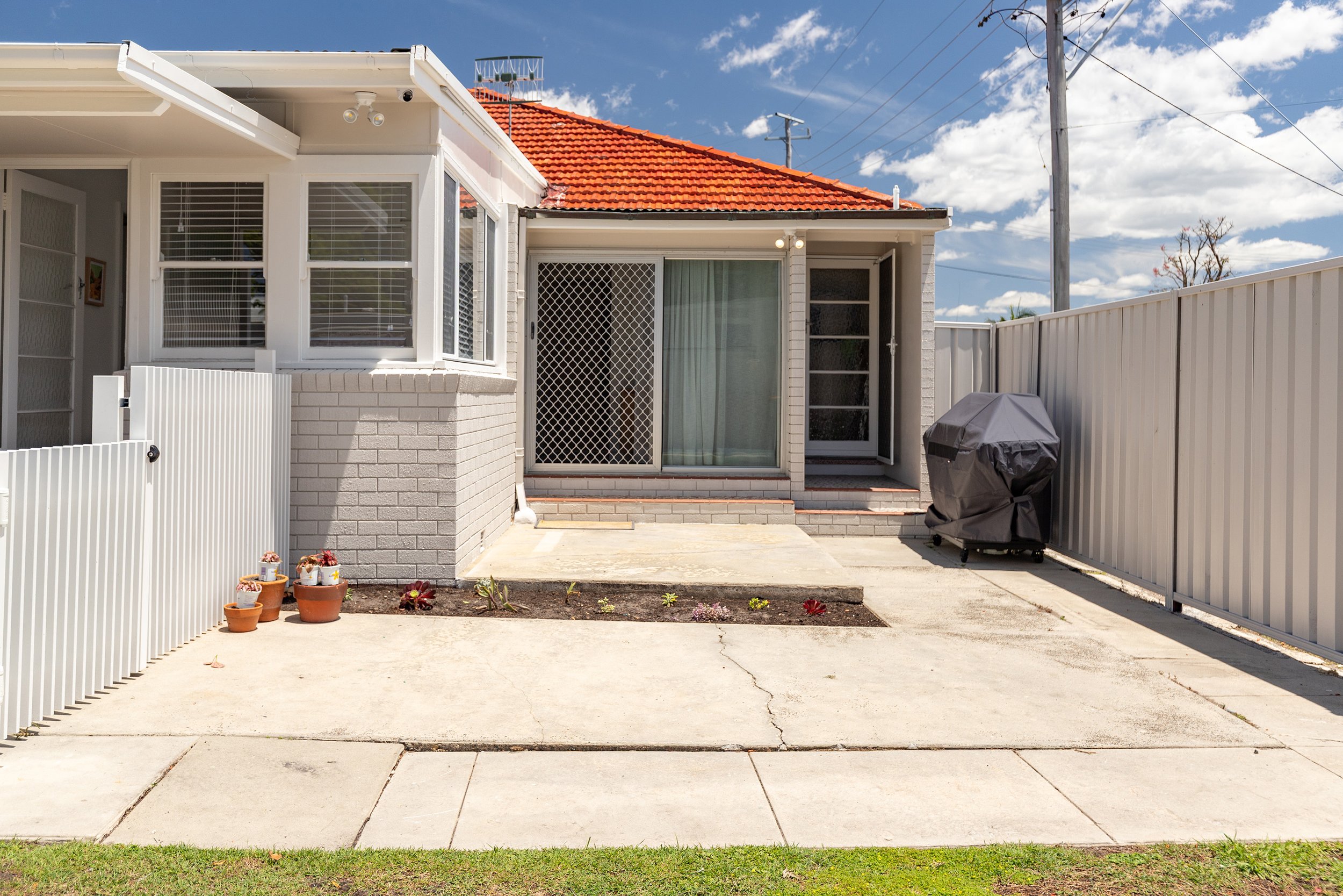 Backyard patio area with concrete walkway, potted plants, small flower bed, sliding glass door with security screen, and grill covered with a black grill cover, enclosed by a white metal fence with utility poles and blue sky with clouds in the backgr