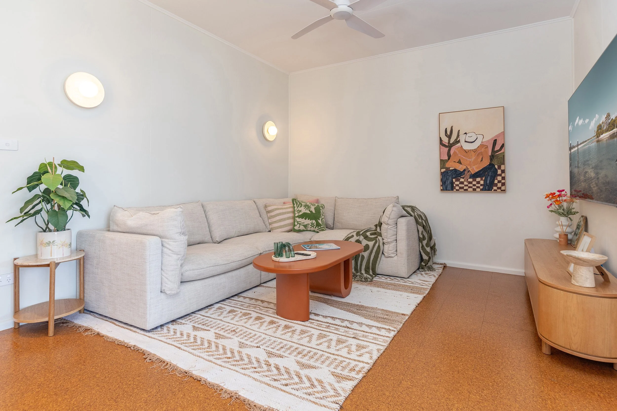 Living room with beige L-shaped sofa, wooden side table with potted plant, orange coffee table, patterned rug, wall-mounted TV, and framed art on the wall.