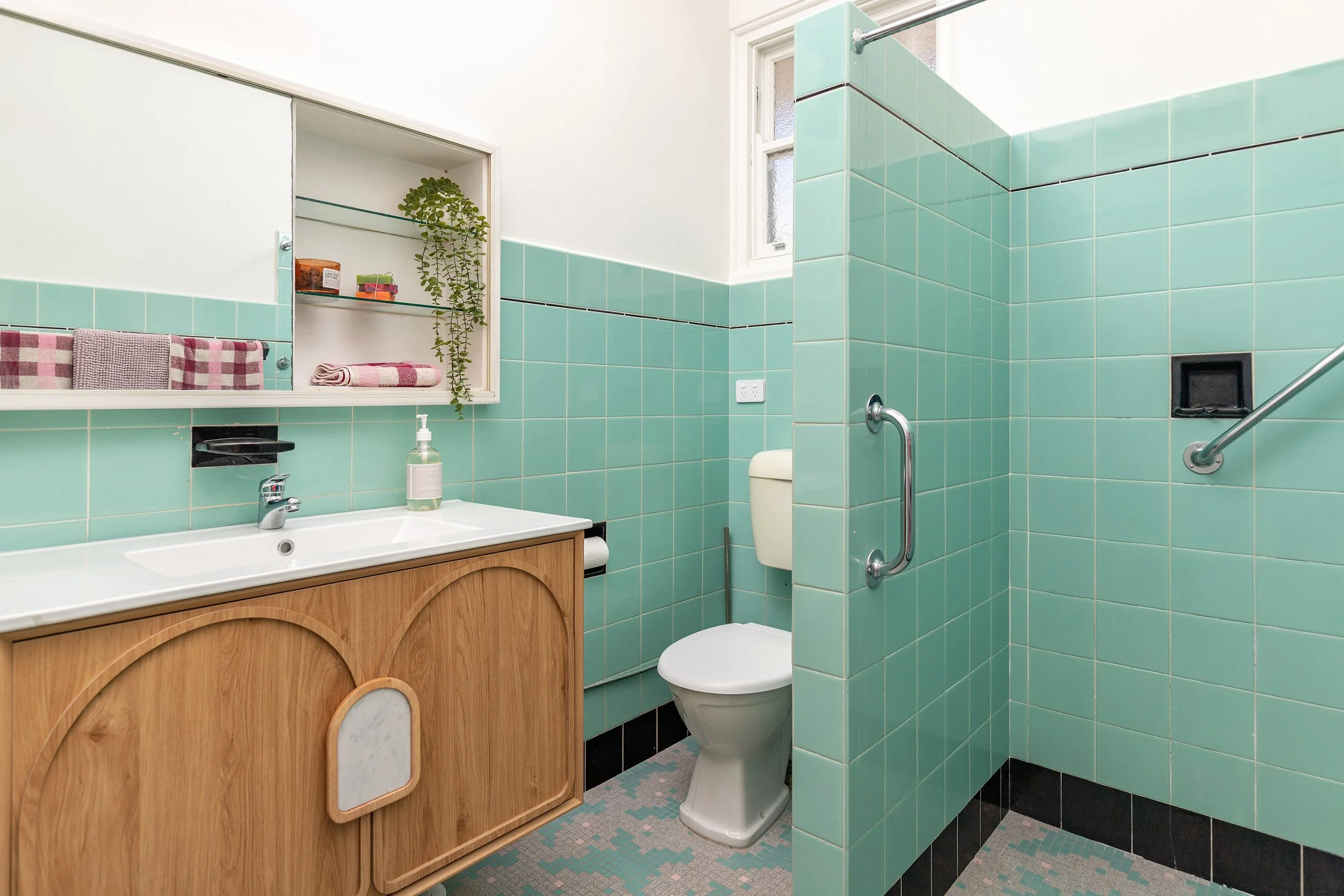 A vintage style bathroom with light aqua tiles, a wooden vanity with a white sink, a toilet, a laundry basket, a mirror cabinet with towels, a soap dispenser, and a small window.