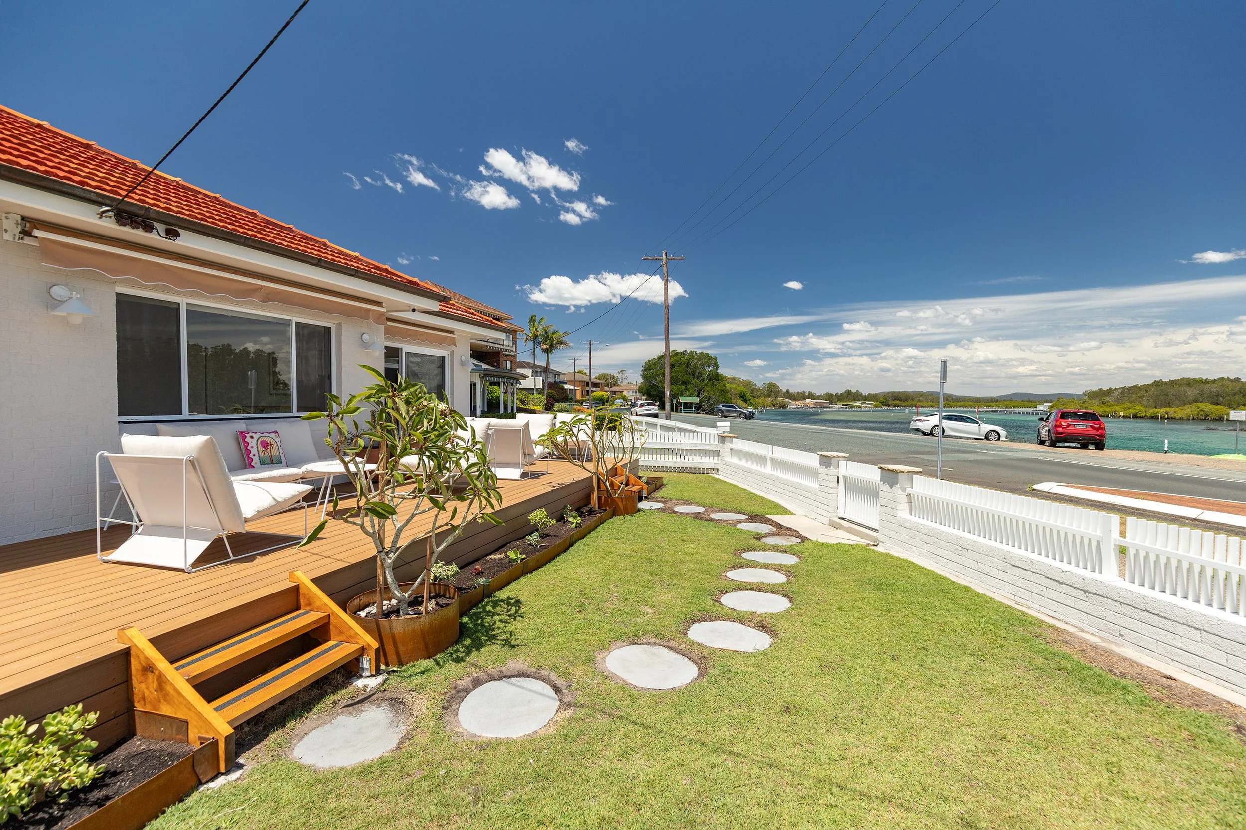 A backyard patio with outdoor seating, a garden, and a view of a waterway with cars parked along the road.