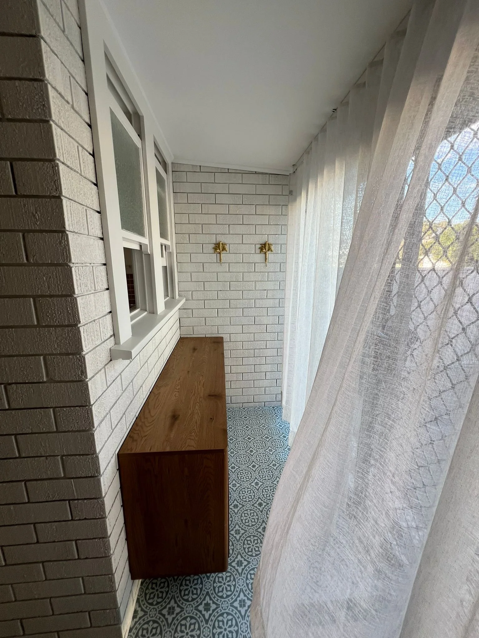 Narrow enclosed balcony with white painted brick walls, a wooden cabinet, decorative patterned floor tiles, sheer curtains, and two gold star-shaped hooks on the wall.