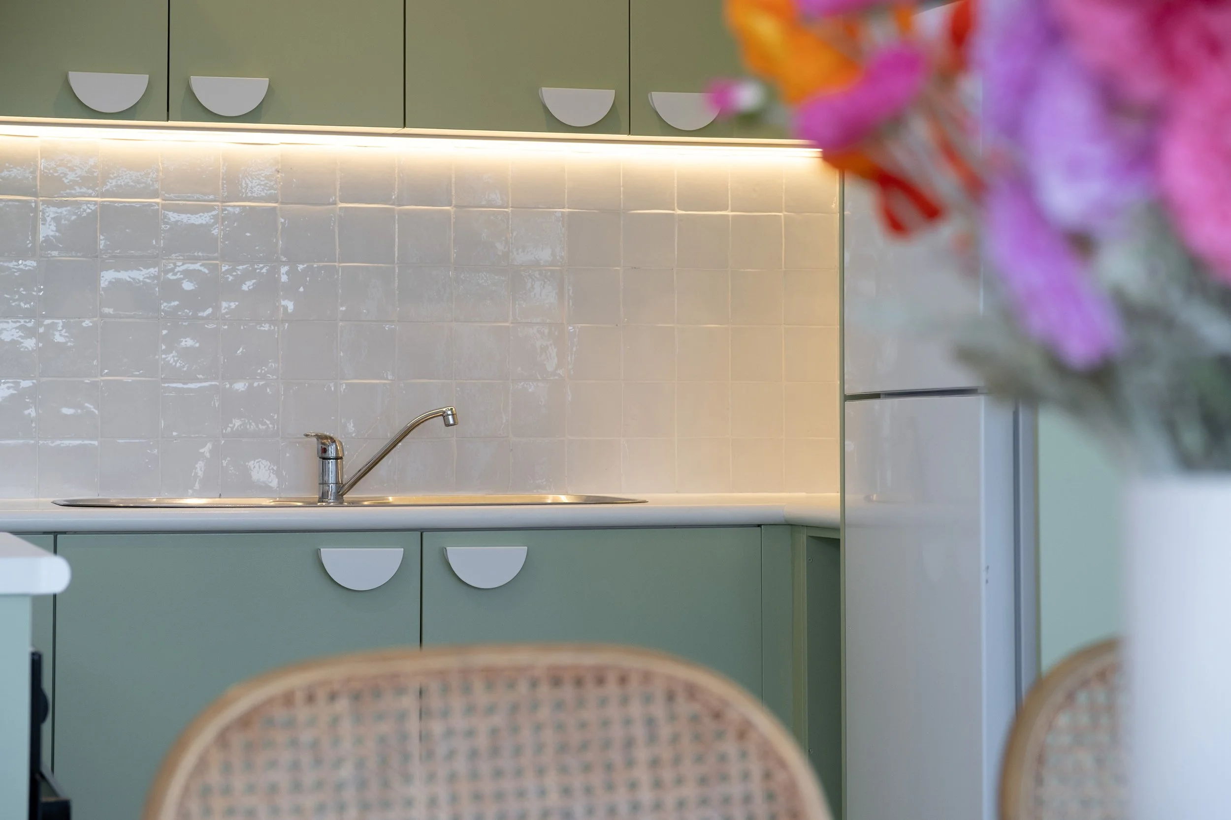 View of a modern kitchen with green cabinets, white tiled backsplash, and a stainless steel sink, with a blurred bouquet of pink and purple flowers in the foreground.