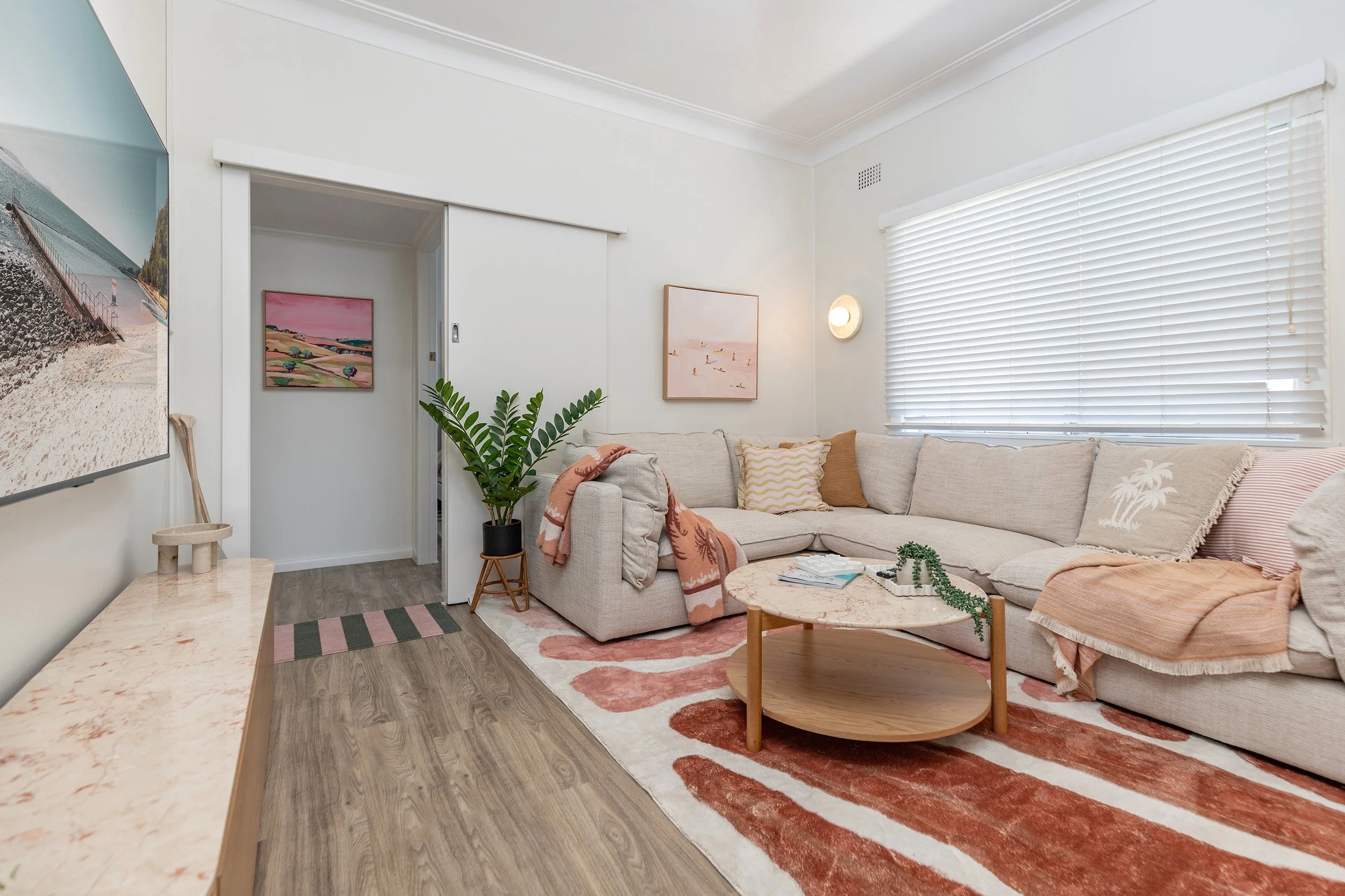 Living room with beige sectional sofa, pink and beige throw blankets, assorted pillows, a round coffee table with books and plants, a patterned rug, a large window with blinds, and wall art. A potted plant is near the doorway.