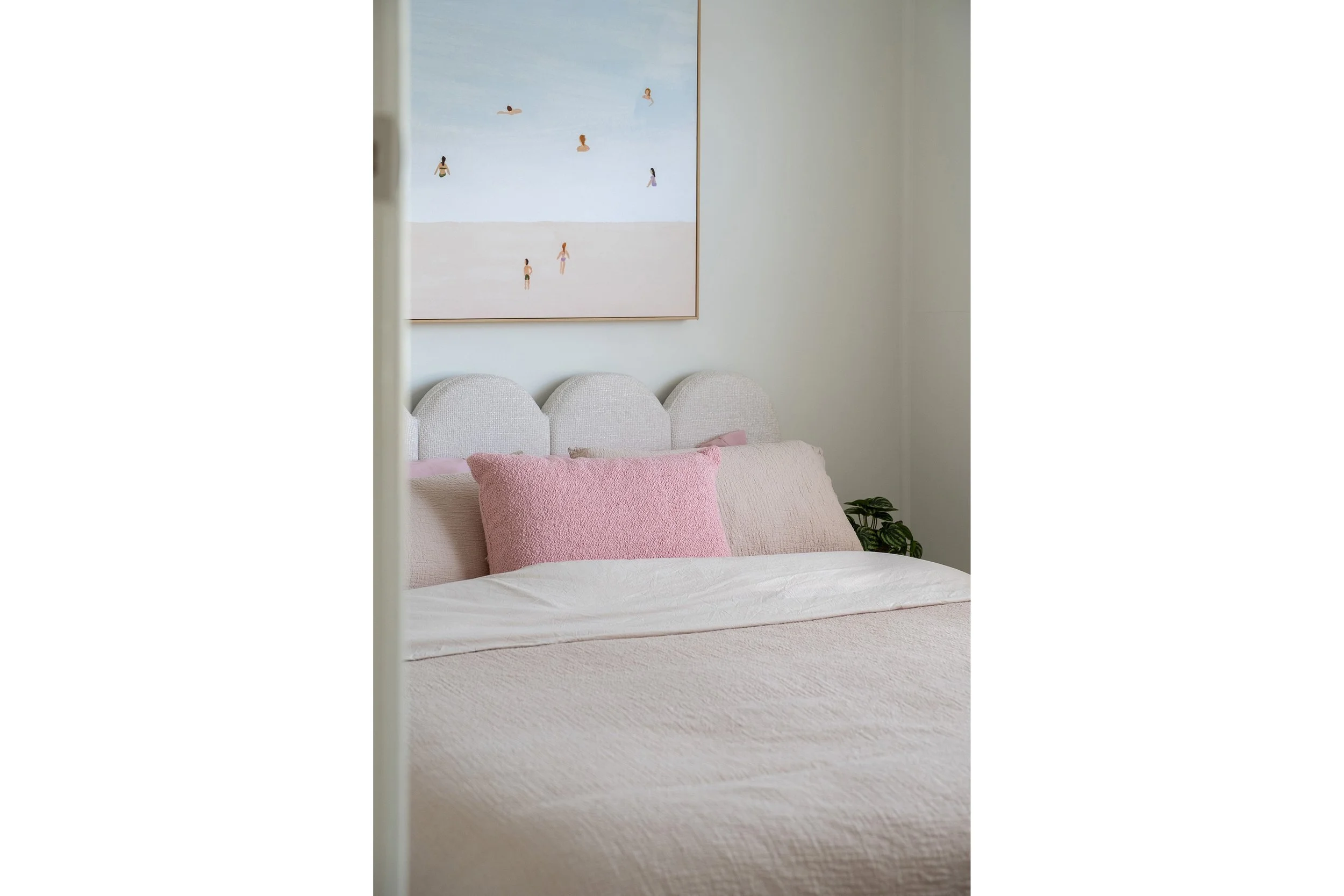 A neatly made bed with soft pink pillows against a white upholstered headboard in a bedroom, with a framed beach-themed art piece on the wall and a green houseplant beside the bed.