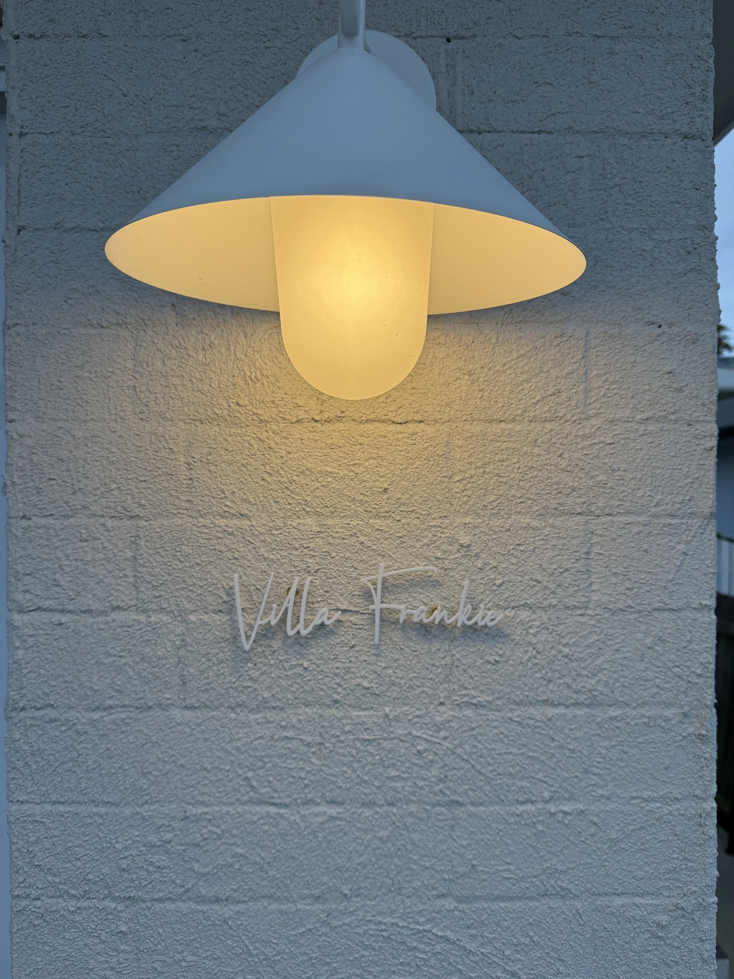 A wall-mounted outdoor light fixture shaped like a flower with a white shade and a frosted bulb, illuminating the wall below. Below the fixture, there is a sign with the text "Villa Frankie" in cursive script.