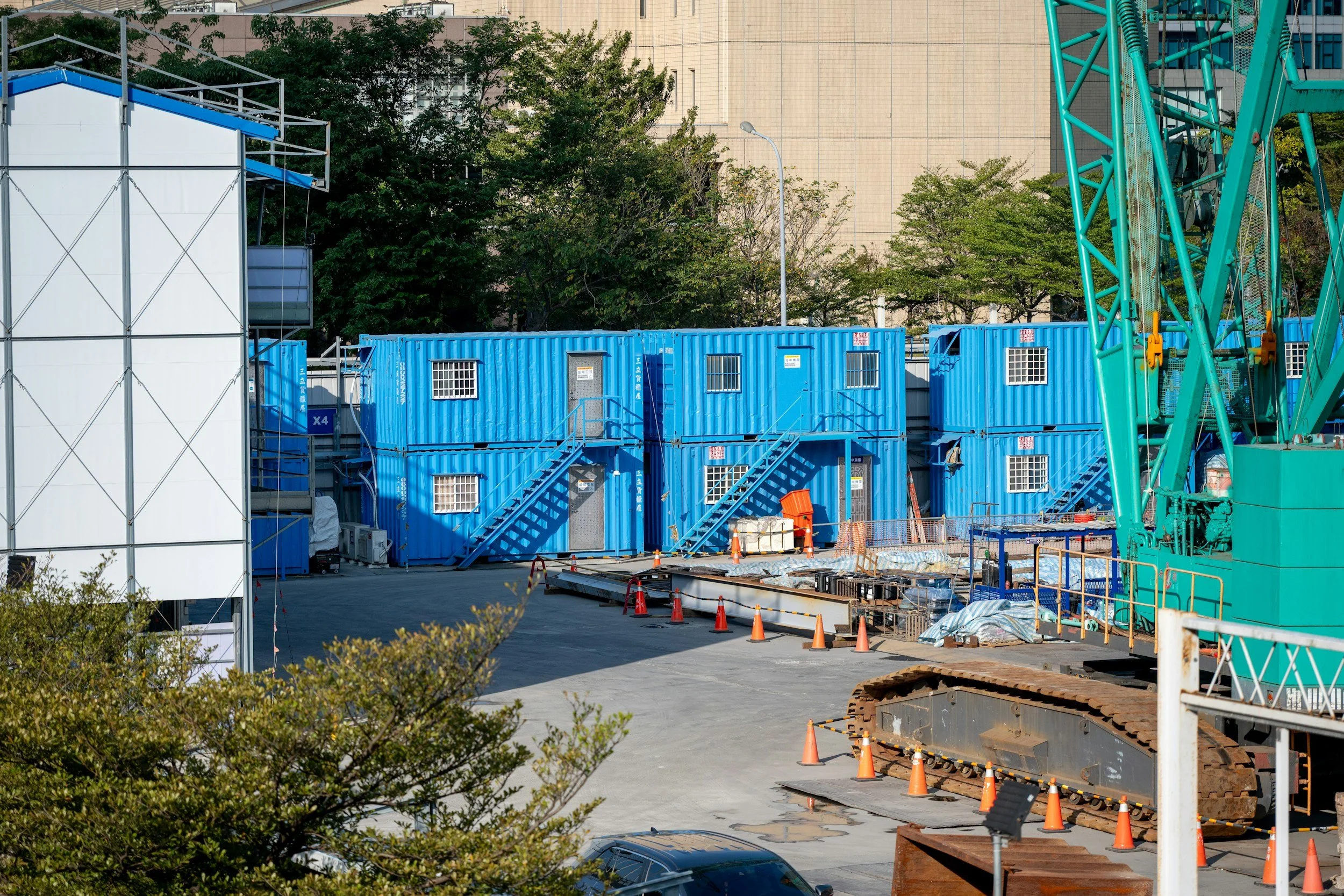 Baustelle mit blauen Container, Bauzaun, Baugeräten und Bäumen im Hintergrund.