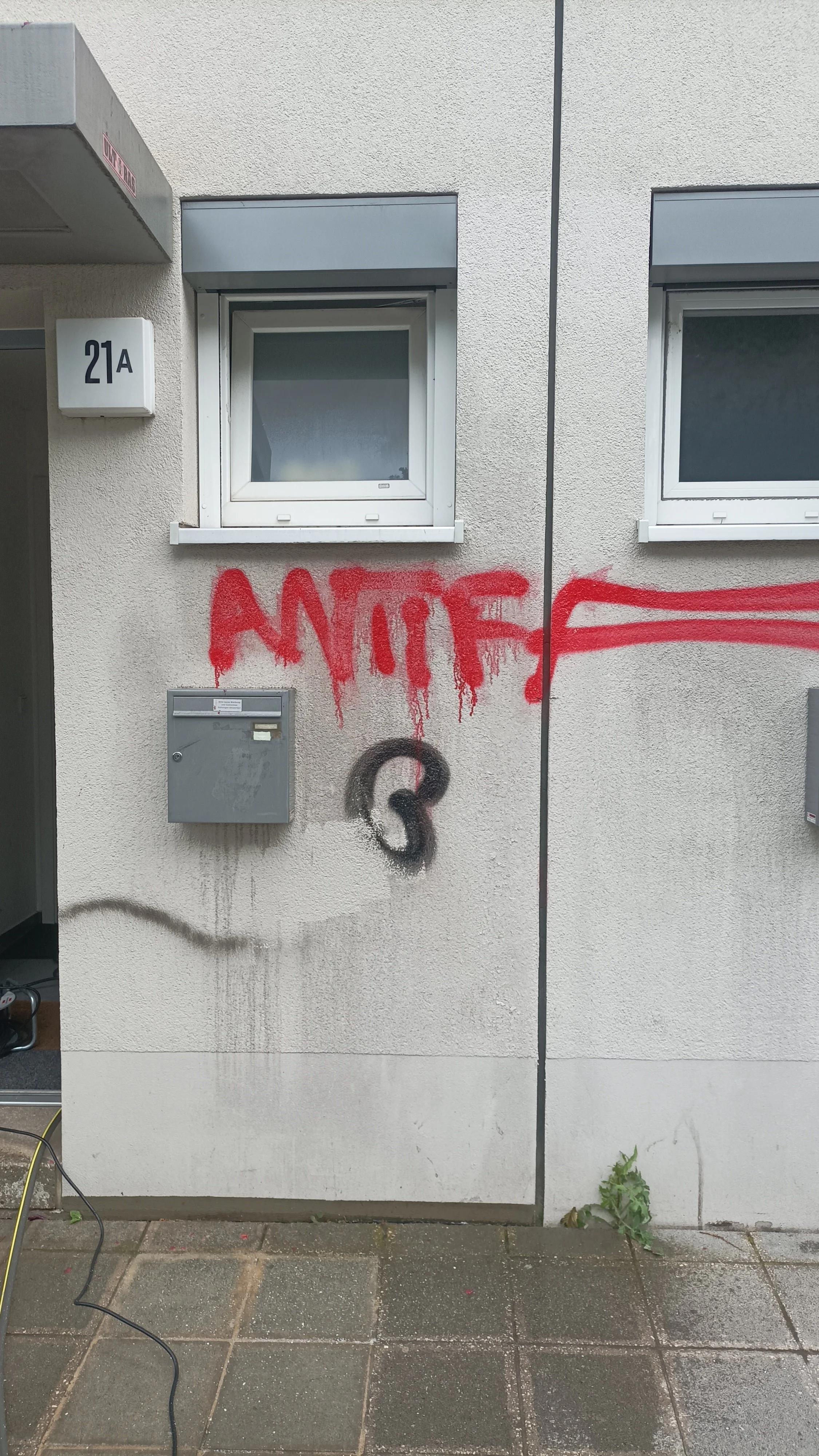 Graffiti in roter Farbe und schwarzer Farbe auf einer Hauswand, daneben ein Fenster und ein Briefkasten.