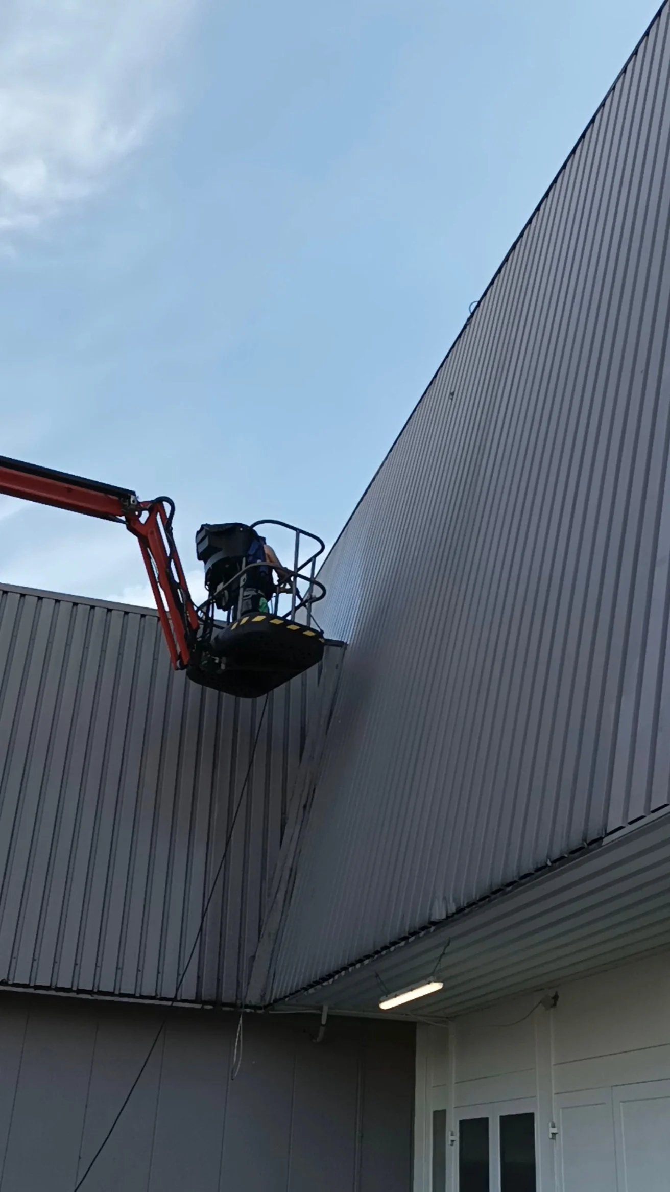 Ein Arbeiter steht auf einem Hubsteiger und arbeitet an der Außenwand eines Gebäudes, das mit Metallblechen verkleidet ist.