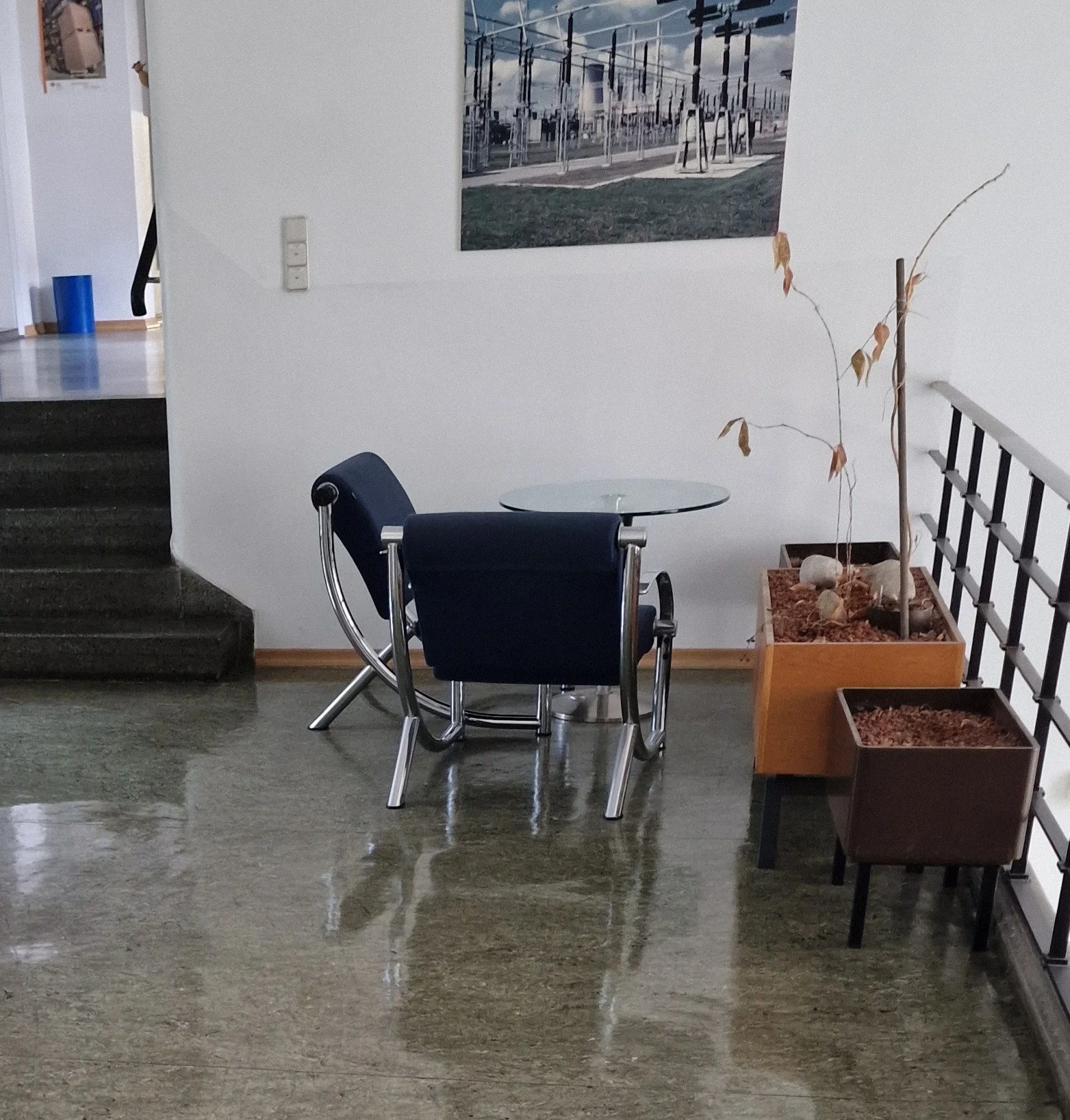 Ein minimalistischer Innenraum mit einem kleinen runden Glastisch, einem modernen schwarzen Stuhl mit Metallgestell, einer dekorativen Pflanze in einem rechteckigen Topf sowie einem Bild an der Wand, das eine Straße mit Stromleitungen und Gebäuden zeigt.