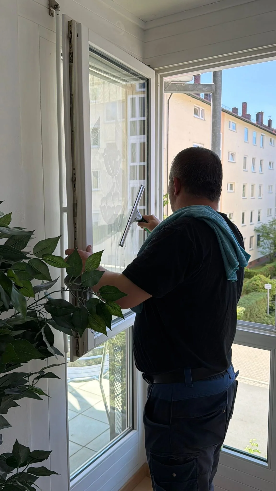 Ein Mann putzt mit einem Fensterwischer ein großes Fenster in einer Wohnung, im Hintergrund sind Wohngebäude und ein blauer Himmel zu sehen.