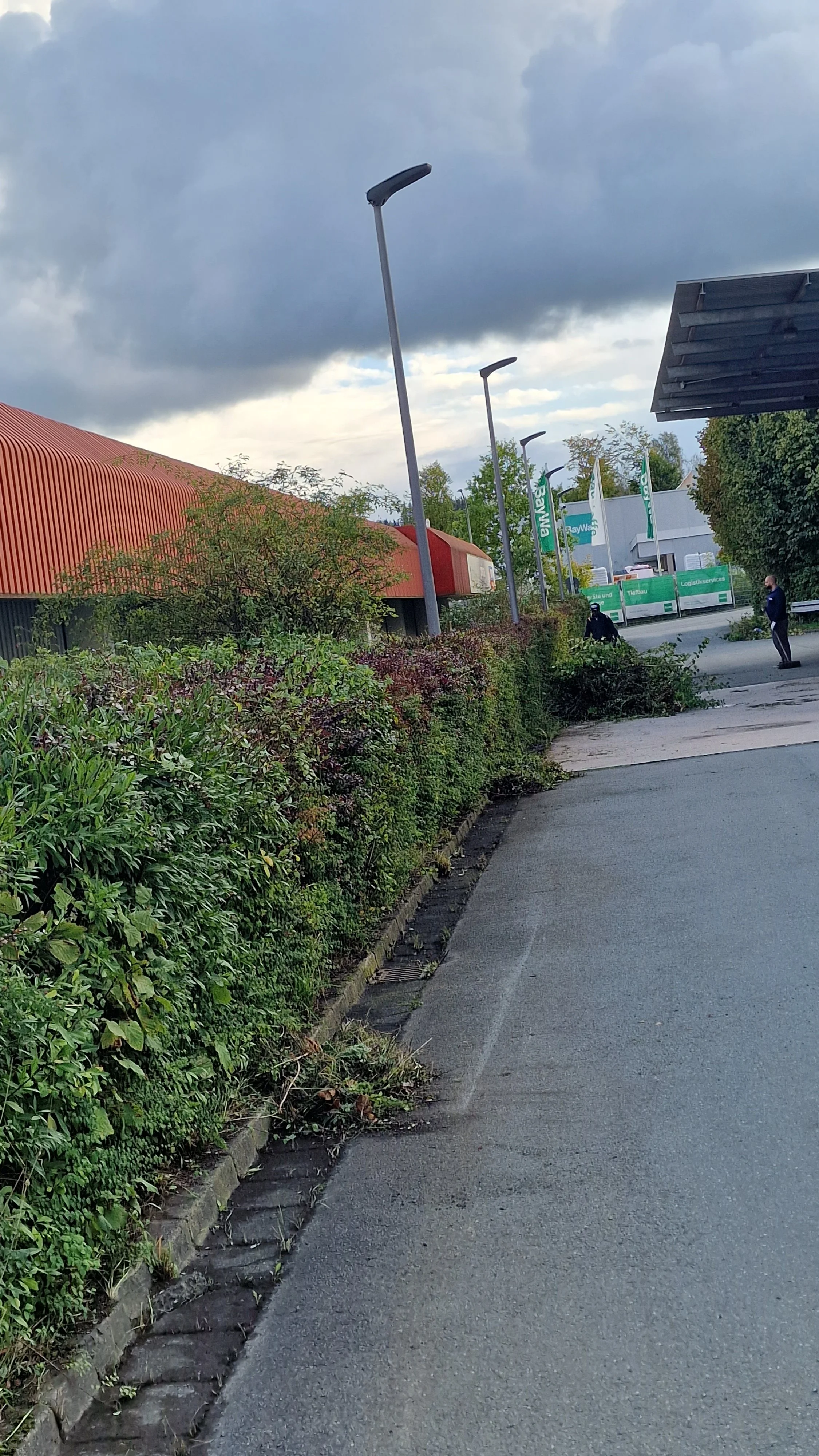 Straße mit Gehweg, auf dem Rasen und Büsche entlang wachsen, überblick mit dunklen Wolken im Himmel, einige Menschen im Hintergrund, Lagerhallen mit grünen Flaggen im Abstand.