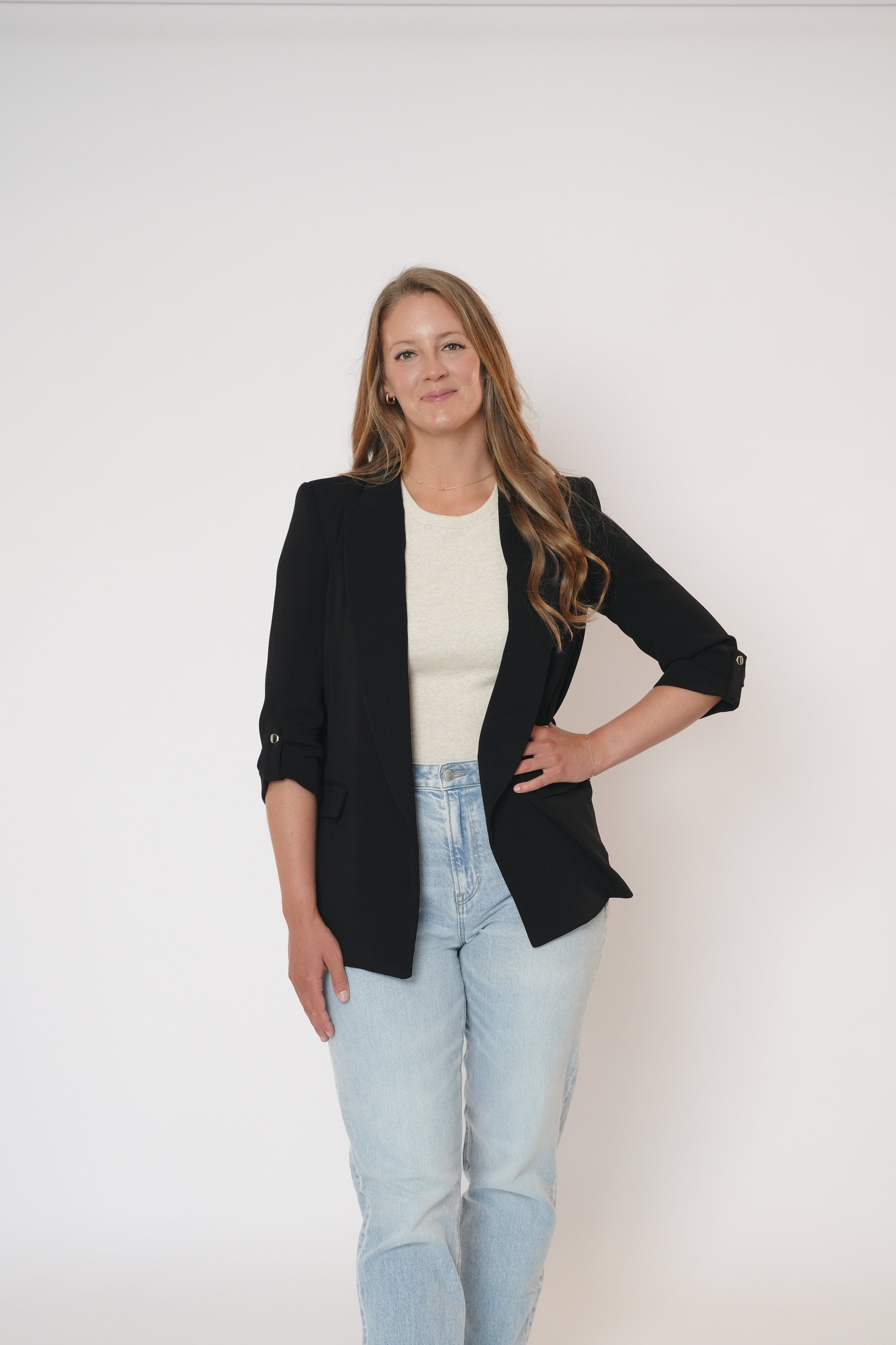 A young woman with long, wavy red hair posing against a plain white background. She is wearing a black blazer, a light-colored top, and light blue jeans.