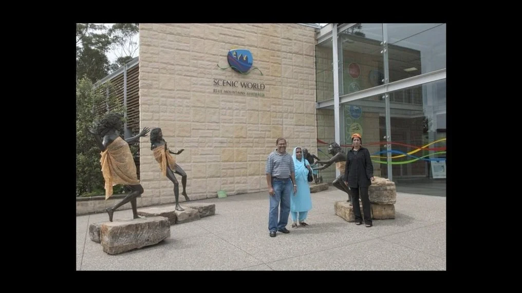 Three people standing outdoors in front of a building labeled 'Scenic World Blue Mountains Australia', posing with four bronze statues of Aboriginal women covered by yellow scarves.