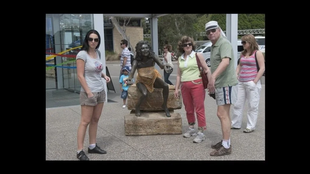Group of people pose straight-faced in front of a bronze sculpture of an Aboriginal woman covered by a yellow scarf.