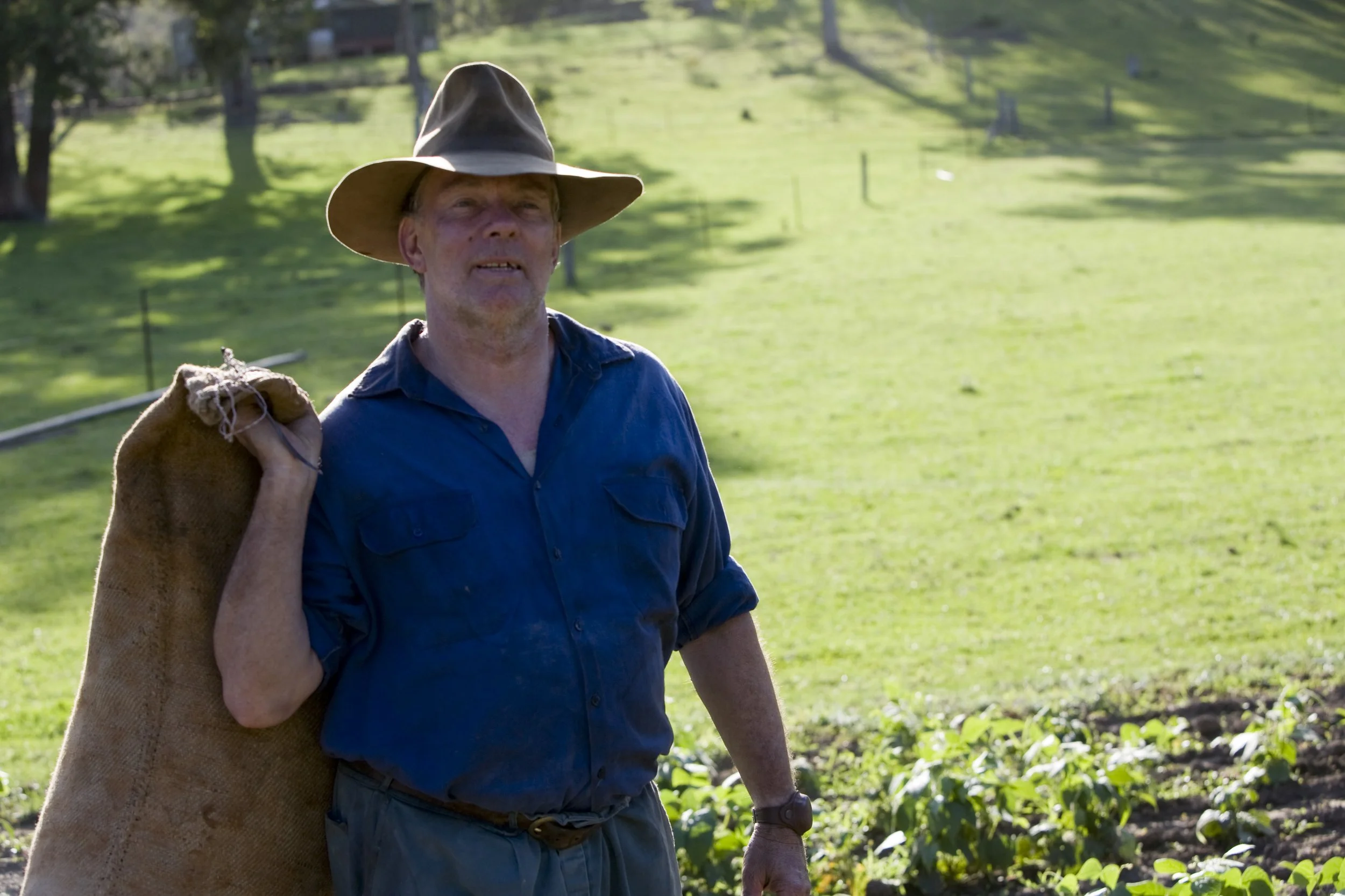 A man wearing a wide-brimmed hat and a blue shirt stands outdoors in a green field, carrying a burlap sack over his shoulder.