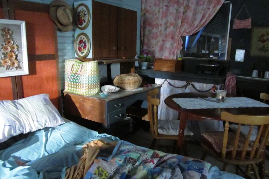 A cozy bedroom with a bed covered in a light blue sheet, a stuffed animal, and a quilt. There are wooden cabinets, decorative plates, and a small desk with bags on it. A round dining table with a lace tablecloth, candles, and wooden chairs is visible