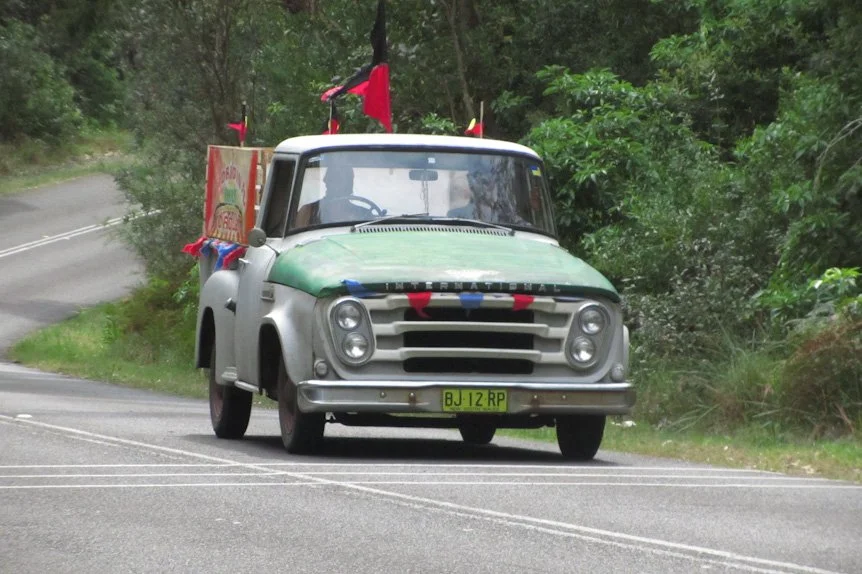 An old Dodge truck decorated with red, white, and blue flags and bunting parked by the roadside surrounded by greenery.