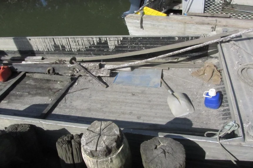 View of an old wooden boat docked on water with various tools and objects on its surface, including a white scoop, blue container, and some wood planks and debris.