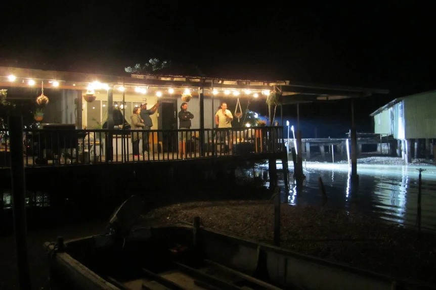 Nighttime scene of people gathered on an outdoor deck with string lights, overlooking a body of water, with reflections of lights and structures on the water's surface.