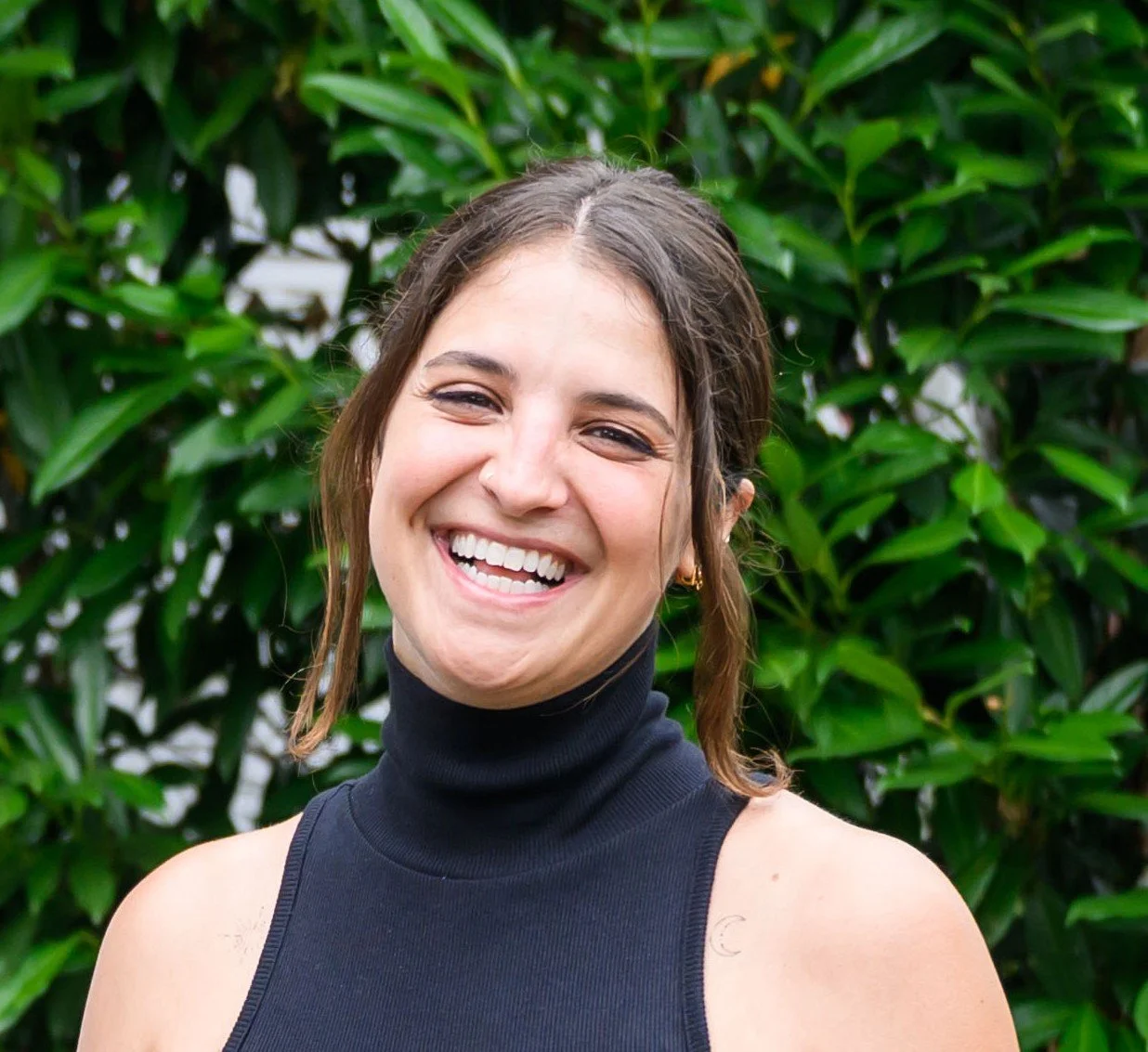 A woman with short brown hair taking a selfie outdoors near a beach, with a cloudy sky, palm trees, and a cityscape in the background, wearing a green jacket and white headphones around her neck.