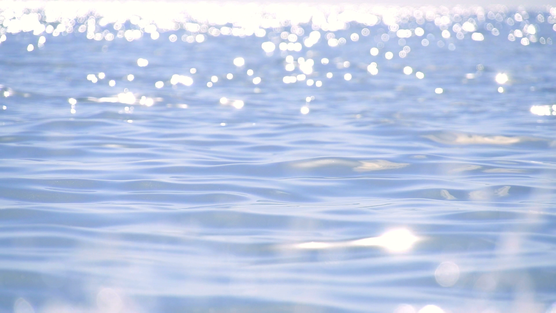 Close-up of calm blue water with sunlight reflecting off the surface, creating bright bokeh effect in the background.