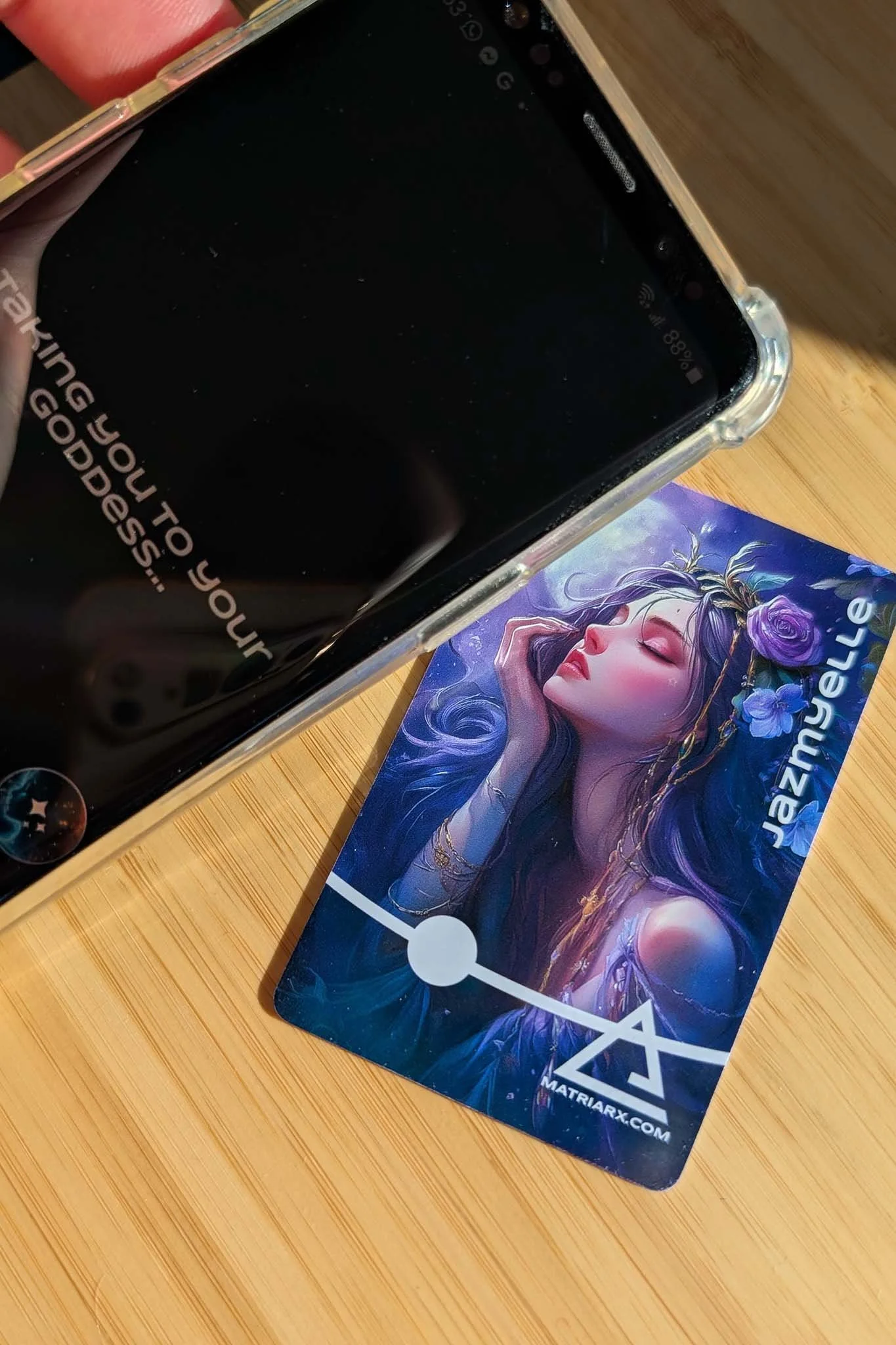 Photograph of a smartphone and a digital art card featuring a fantasy woman with long purple hair, closed eyes, and flowers in her hair, placed on a wooden surface.
