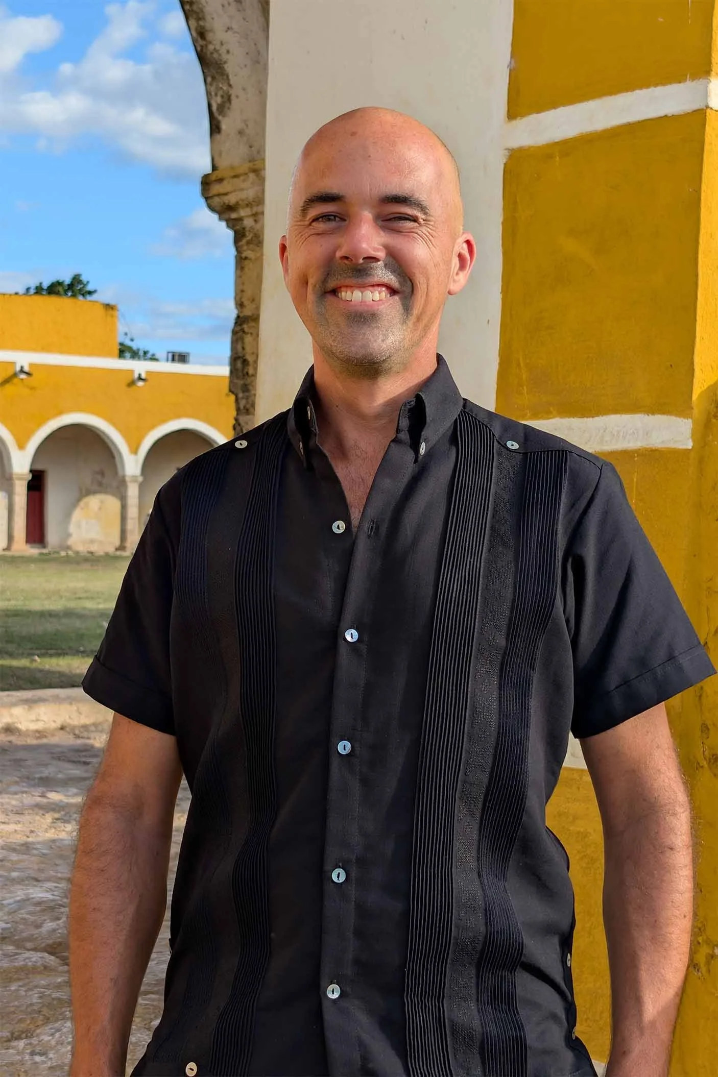 A bald man in a black shirt standing outdoors near a yellow building with arches, smiling at the camera.