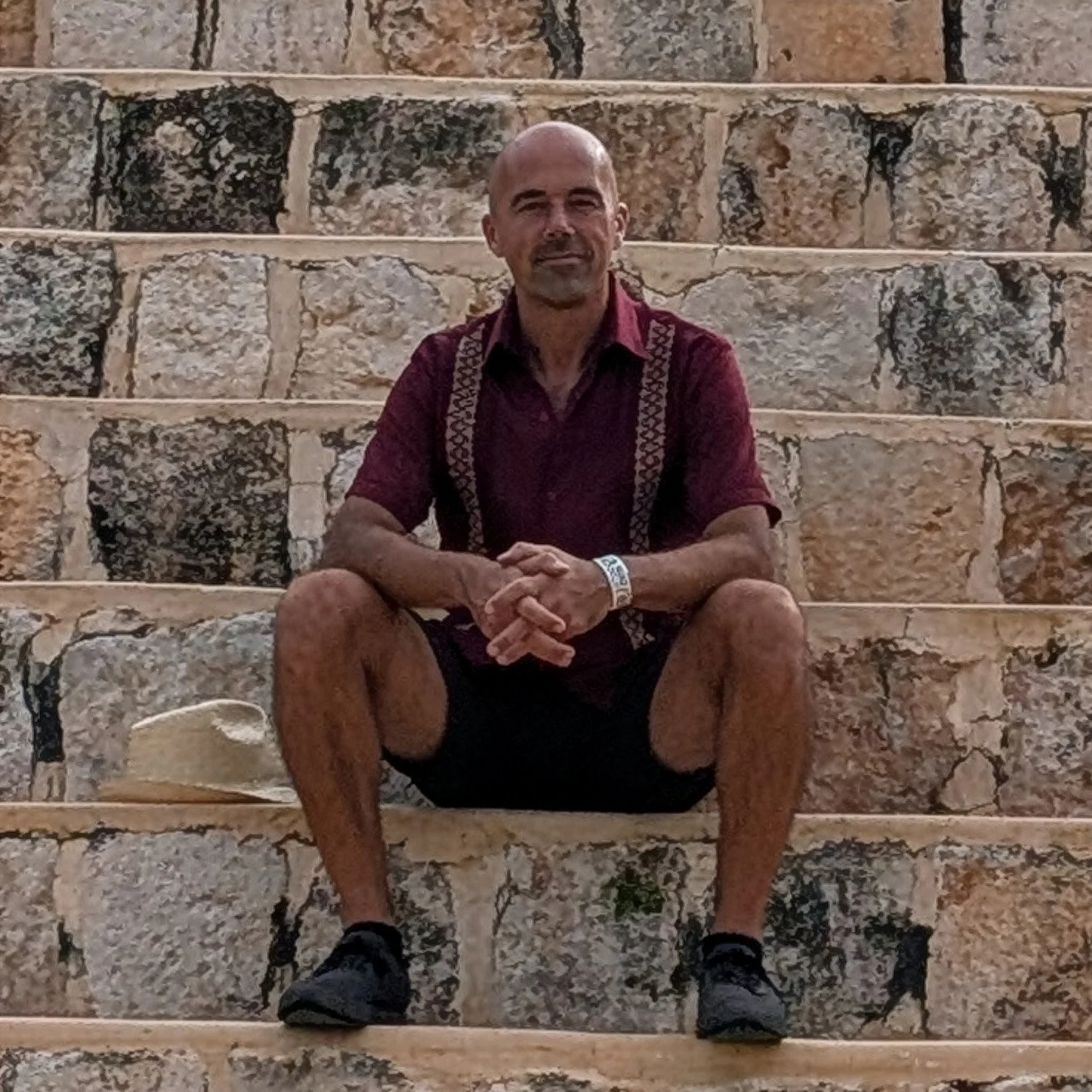 A man sitting on stone steps with his legs spread apart, wearing a burgundy short-sleeve shirt, black shorts, black athletic shoes, a white wristband, and a patterned strap around his shoulders, with a beige hat on the step beside him.