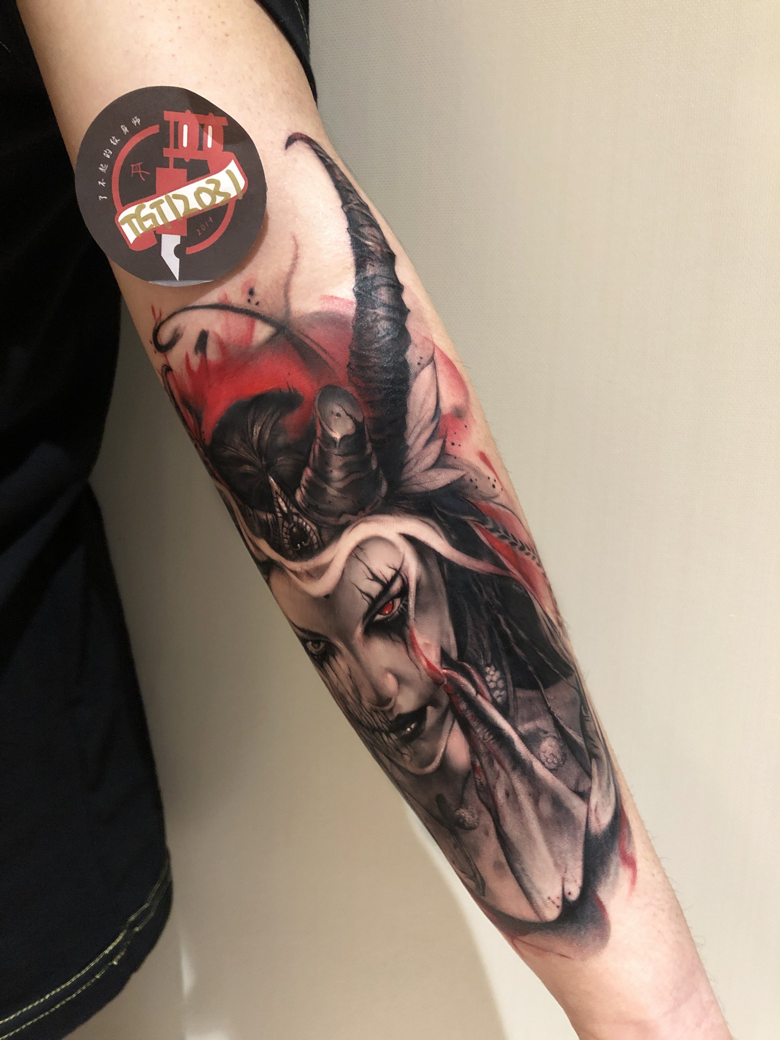 Forearm tattoo with a demonic figure, featuring horns, red accents, and intricate details. A circular sticker with text is near the elbow.