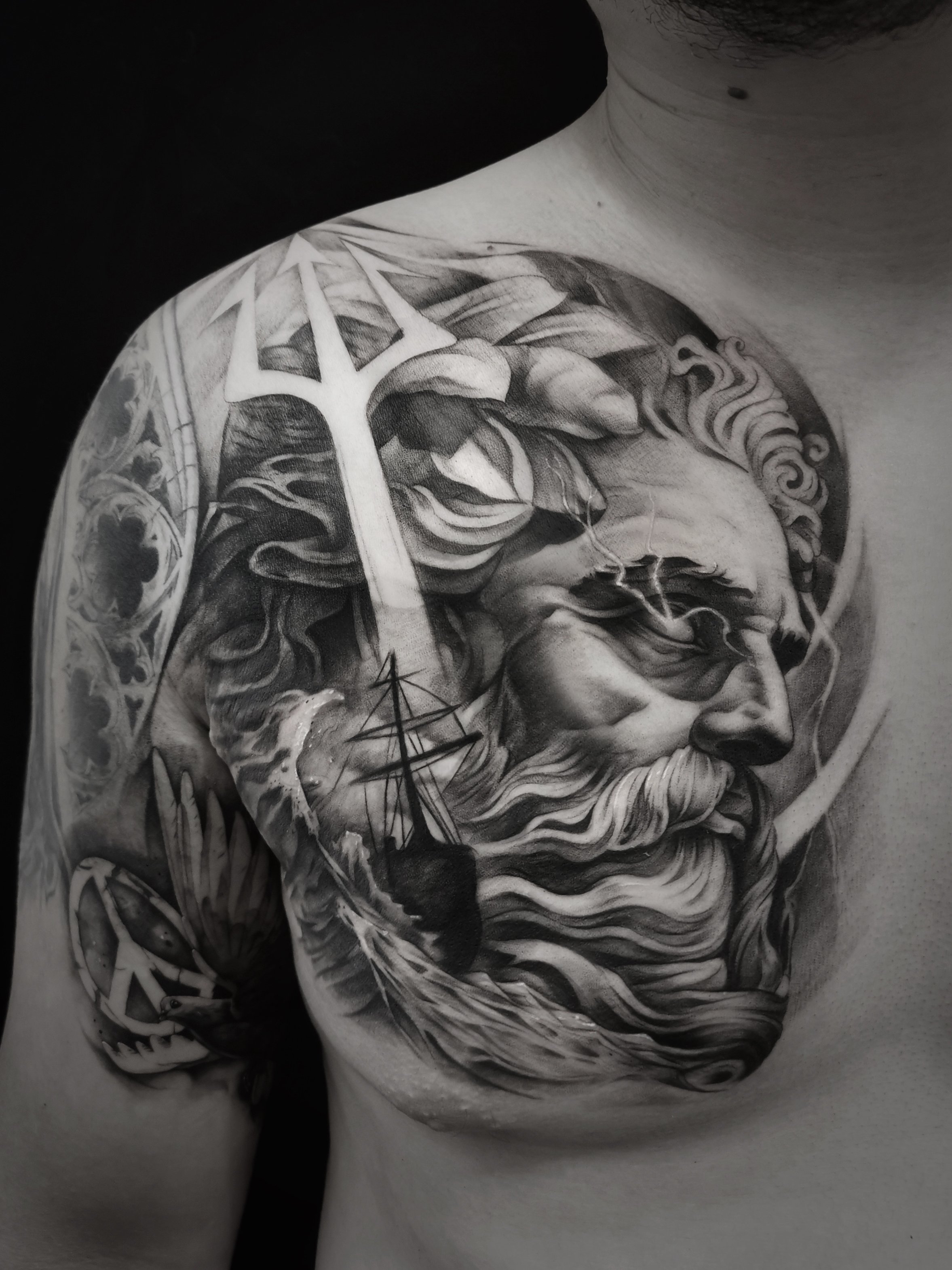 Black and white tattoo of a mythological god with a trident, waves, and a ship on a person's chest and shoulder.
