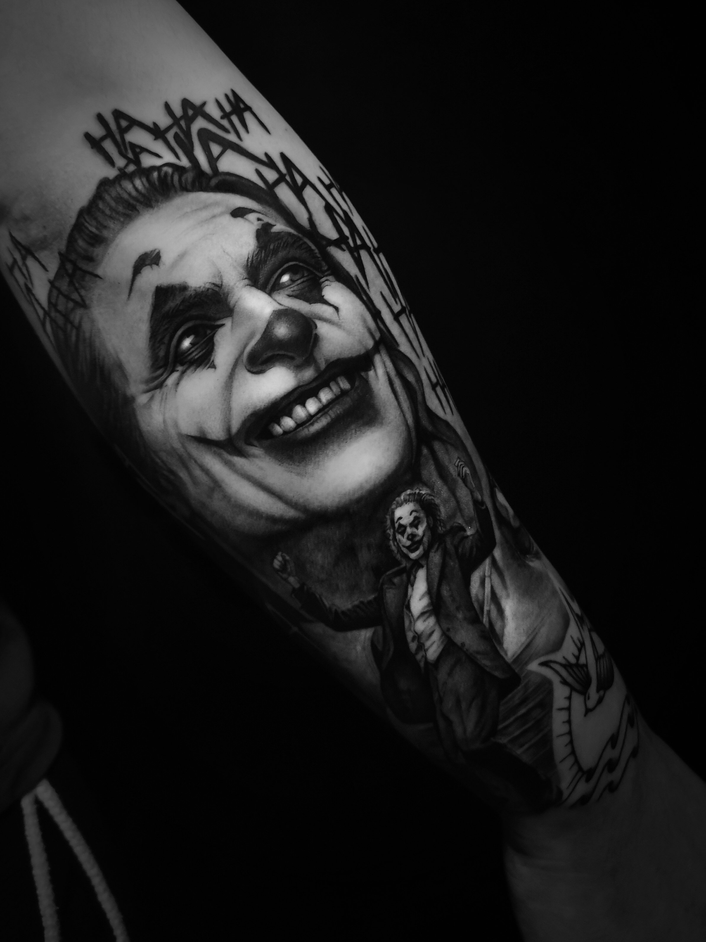 Black and gray tattoo featuring a clown-like face with a wide smile and crazy laughter text. Below, a figure in a suit is depicted, arms raised, against a dark background. The overall mood is intense and dramatic.