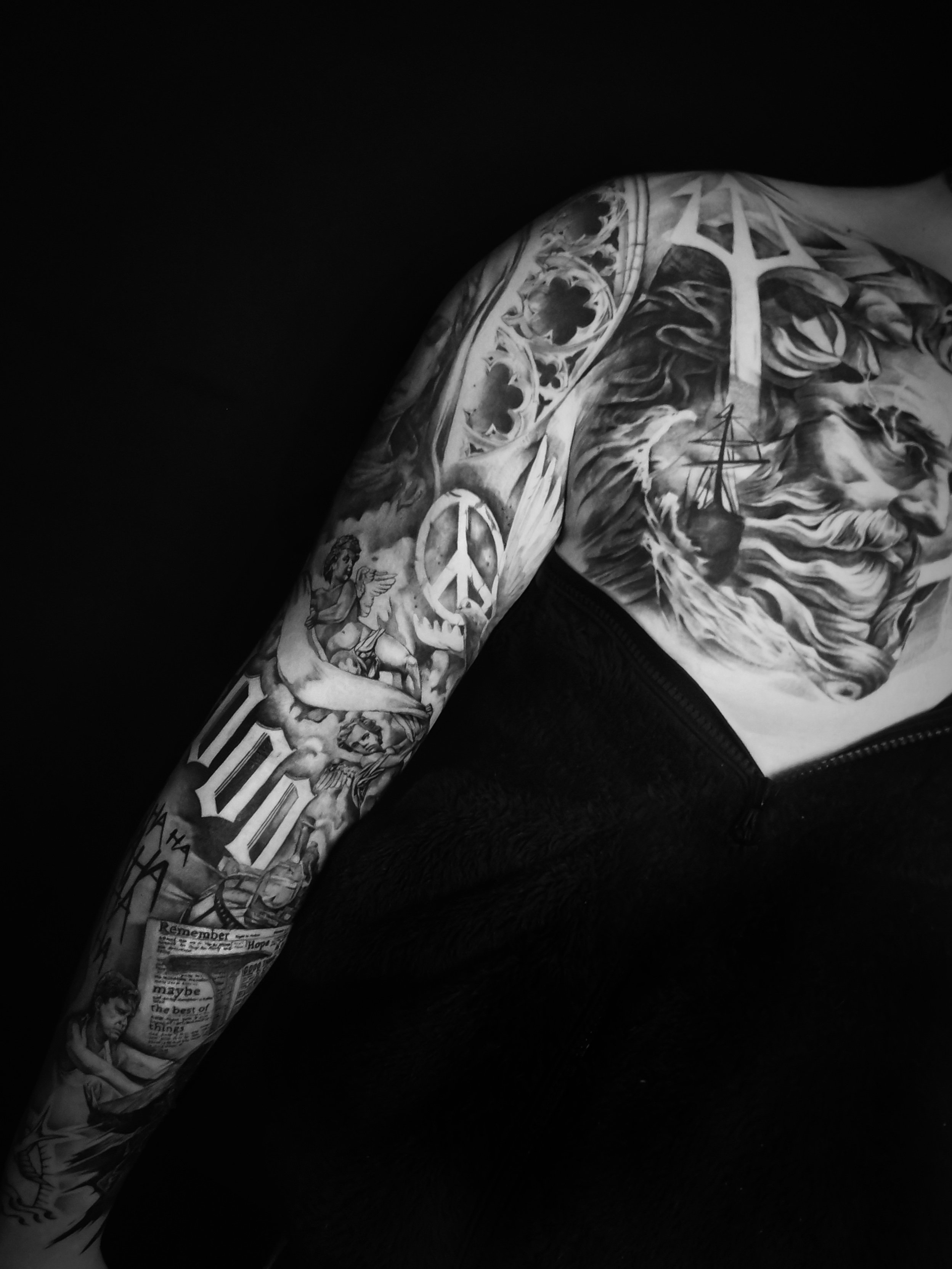 Black and white tattoo sleeve with various intricate designs, including a lion, peace symbol, angels, gothic architecture patterns, newspaper clippings, and other artistic elements.