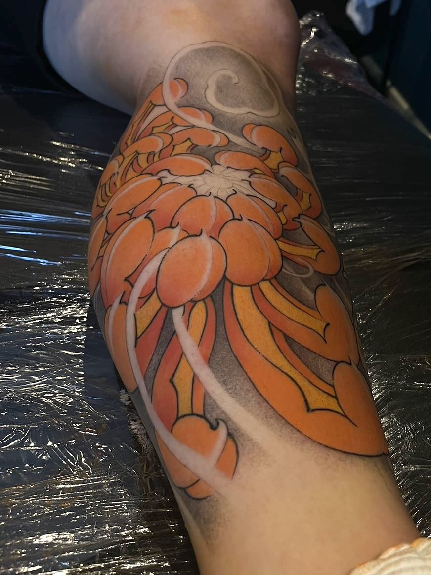 Orange and black tattoo of a chrysanthemum on a person's calf.