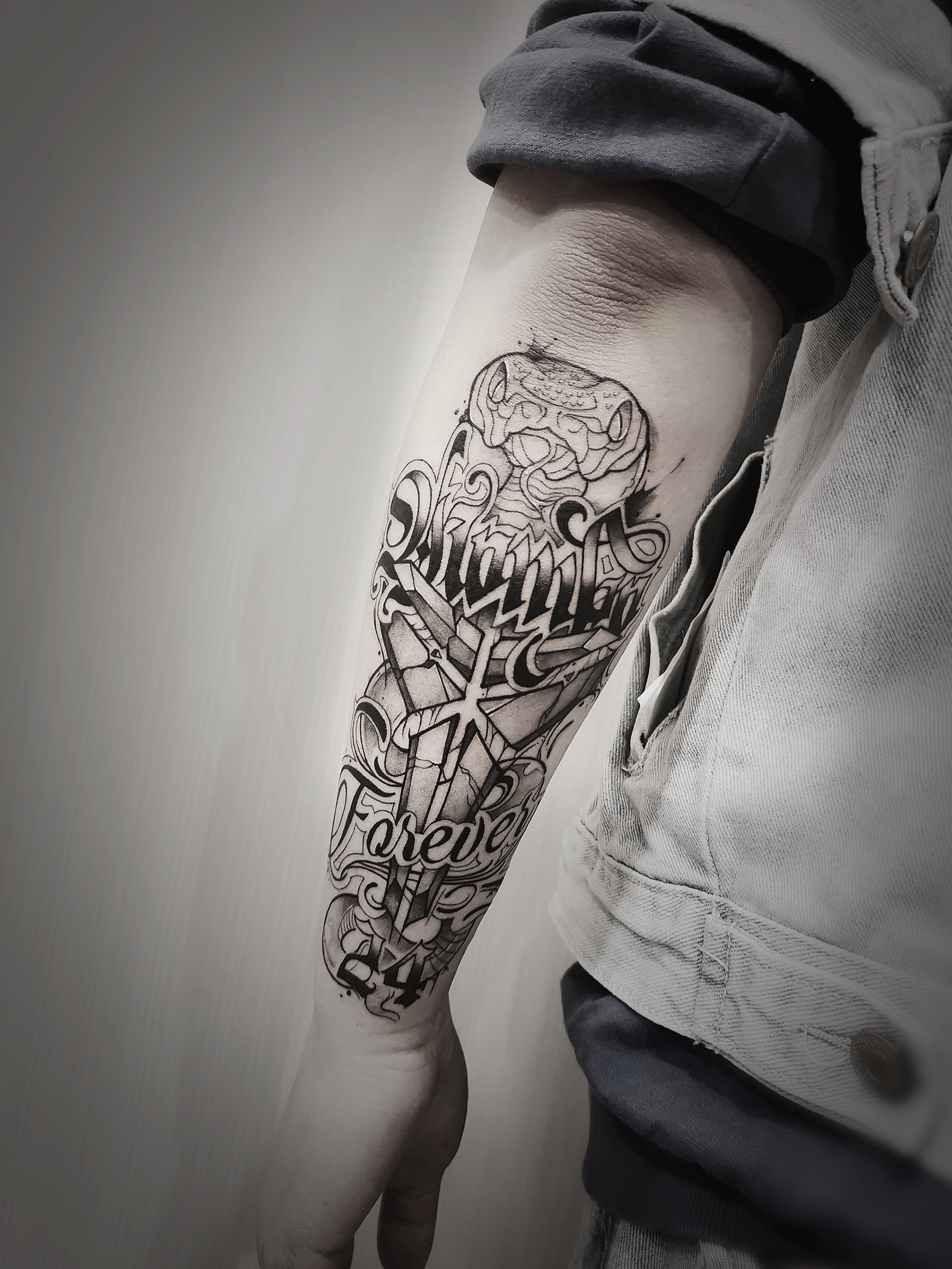 Forearm tattoo featuring a stylized snake, intricate lettering, and a cross design with the words "Forever" and "24" in black ink.