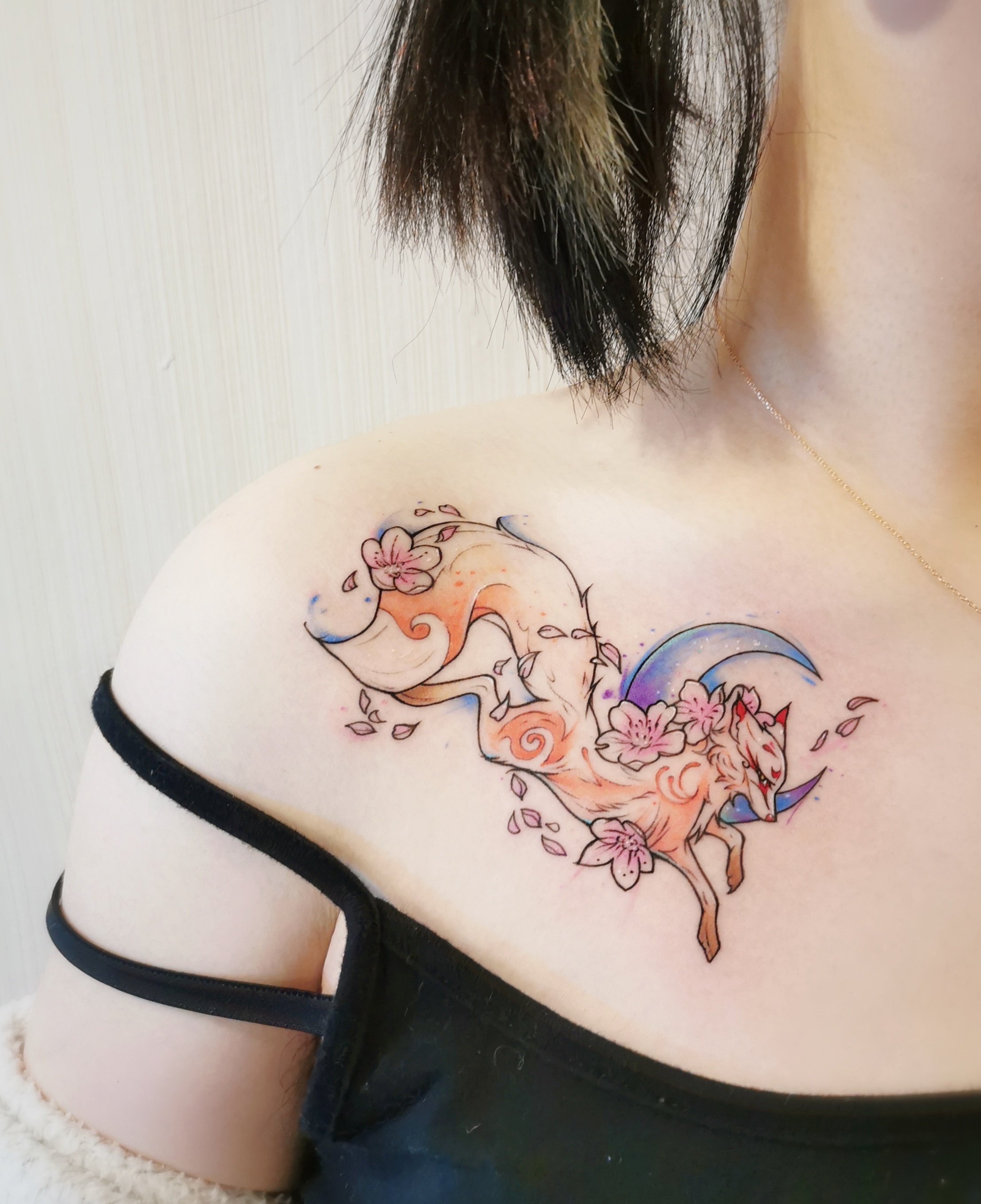 A colorful tattoo on a person's shoulder depicting a stylized fox with floral elements and a crescent moon.