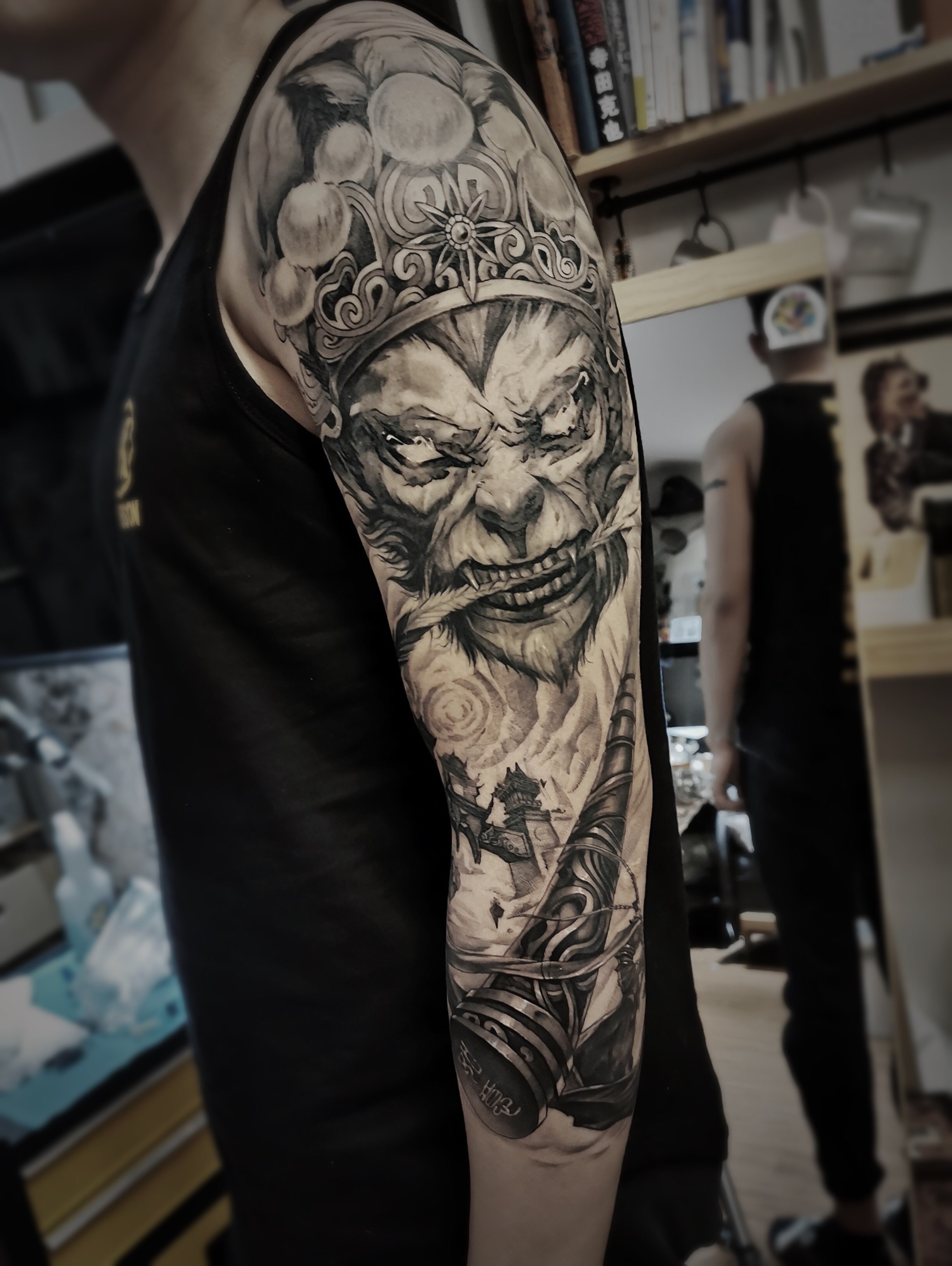 Detailed black and gray tattoo on a person's upper arm, depicting a fierce face wearing an ornate crown, holding a long pipe, with additional intricate patterns and designs extending down the arm.