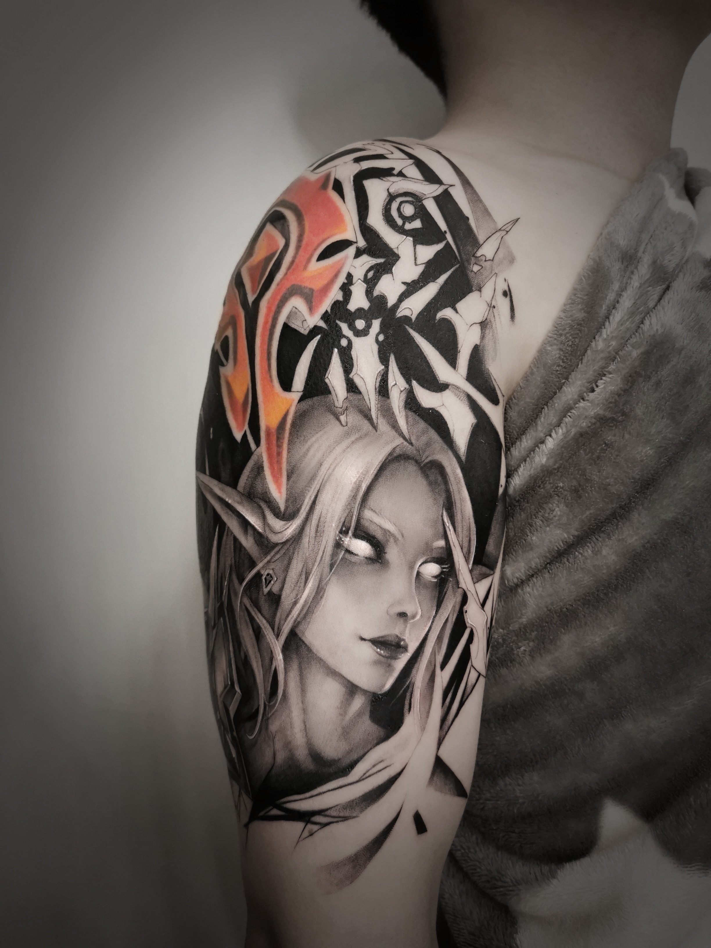 Arm tattoo featuring a fantasy portrait of an elf with long hair and glowing eyes, combined with abstract black and white designs, and a red and orange emblem resembling a tribal symbol on the shoulder.