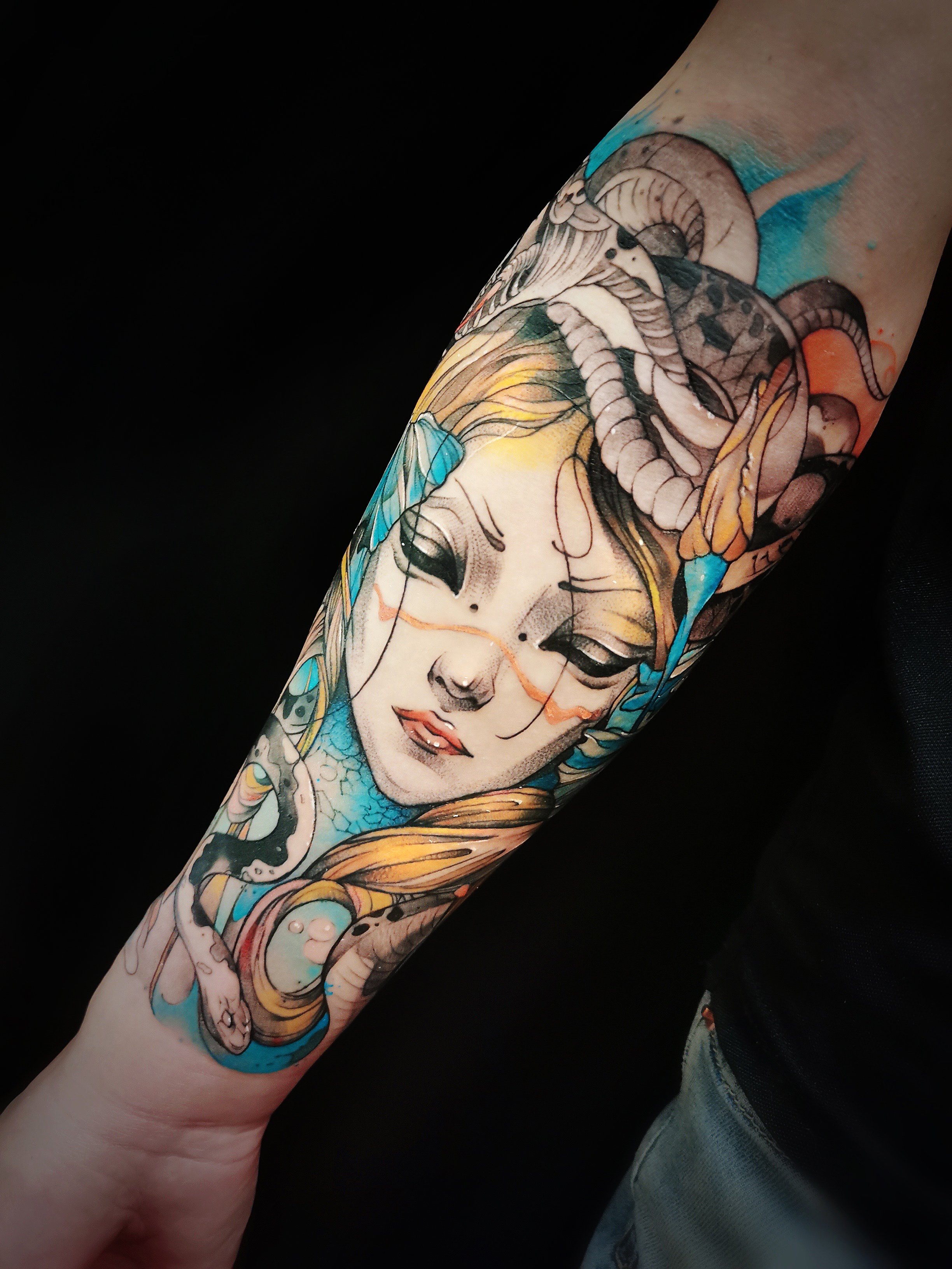 Colorful tattoo on forearm depicting a woman's face with elements like snakes, a blue butterfly, and abstract details.