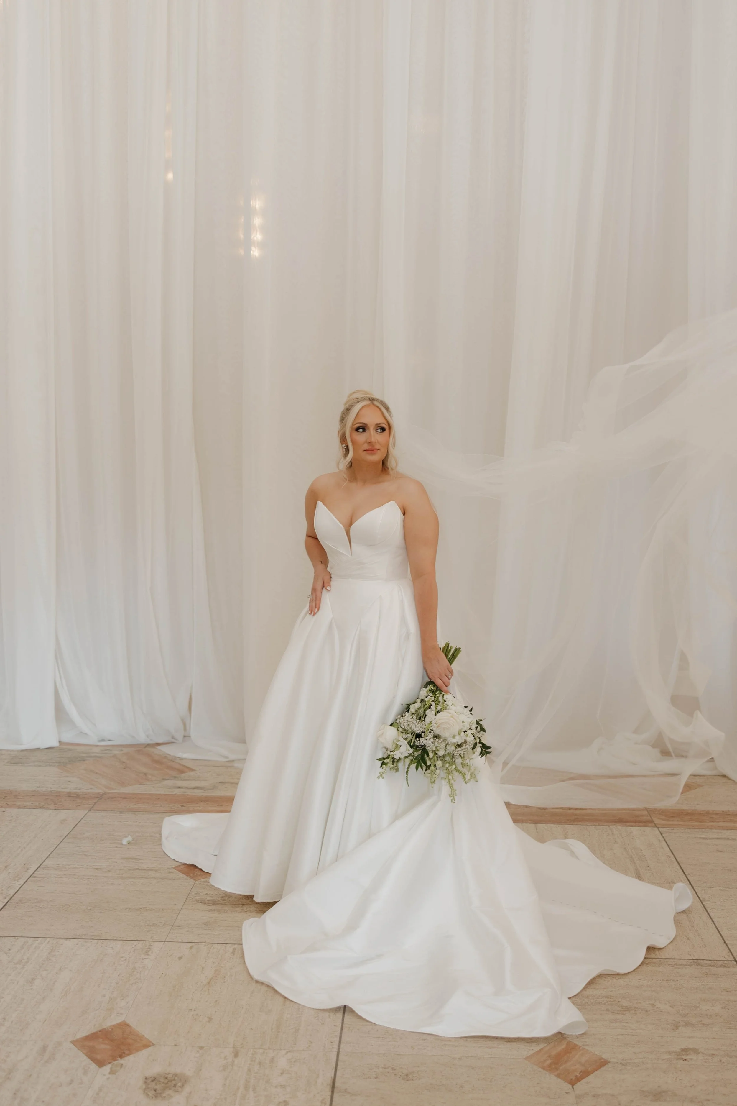 A bride stands in front of ivory drapery holding a bouquet of greenery and white flowers. She is wearing a ballgown and her veil is cascading off to her side.