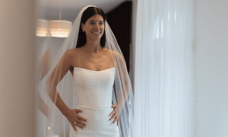 Bride smiling in a strapless white wedding dress and veil, standing indoors with hands on hips. Soft lighting and sheer curtains create a serene atmosphere.