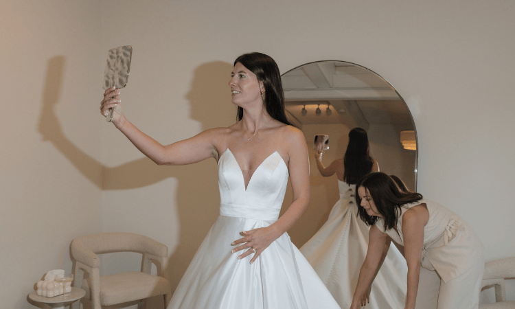 A smiling bride in a white gown holds a mirror, admiring her reflection. A woman helps adjust the dress. The setting is tranquil and joyful.