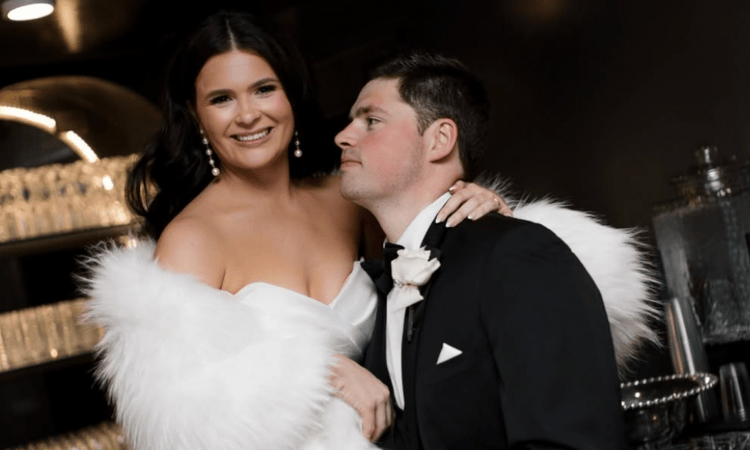 A bride in a white dress and feather shawl smiles warmly, leaning on the groom in a black tuxedo. The setting is elegant and softly lit, conveying joy.