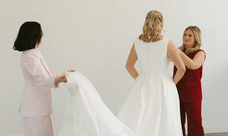 Bride trying on a structured white wedding gown while two bridal stylists adjust the train during a boutique appointment