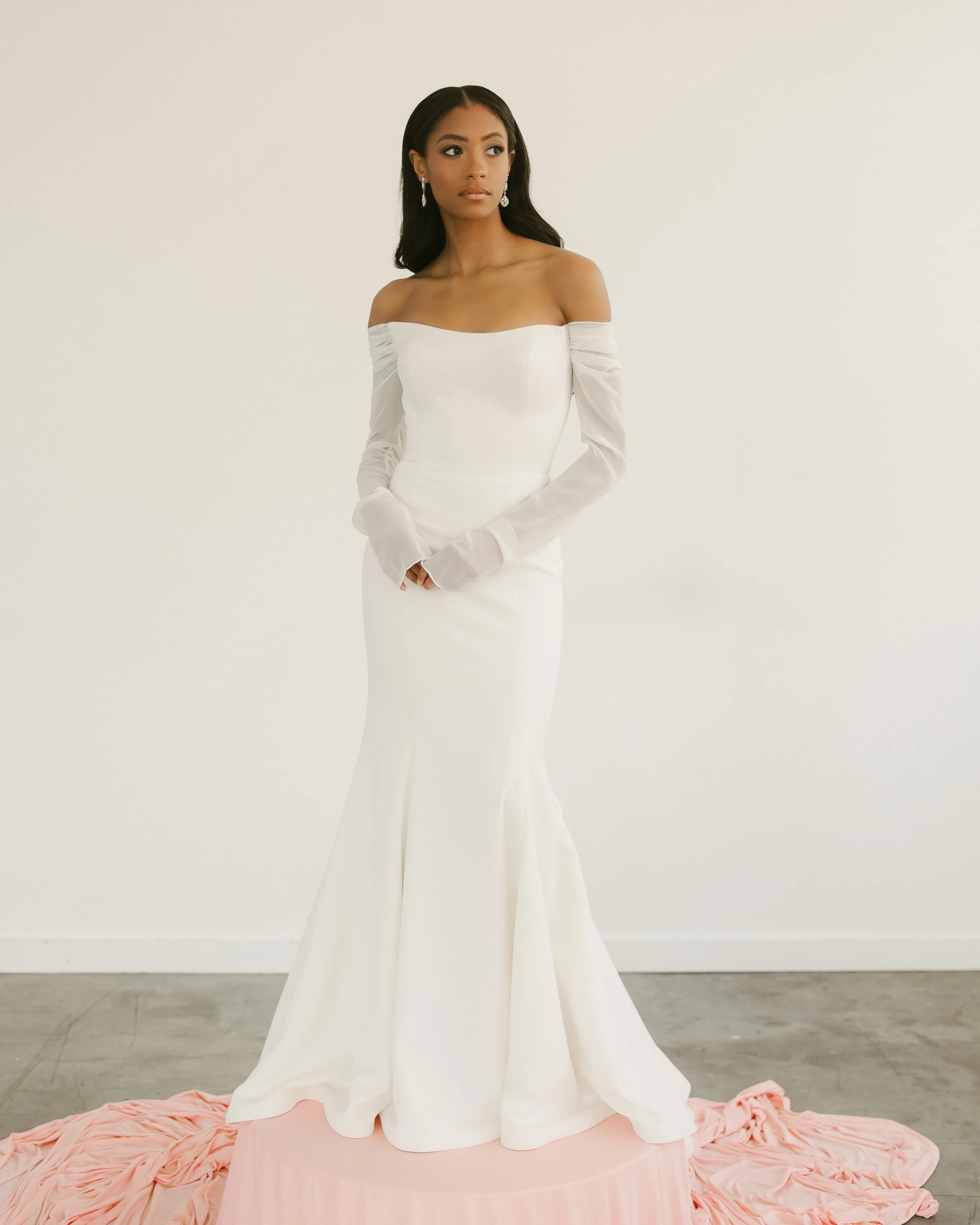 A woman stands in a white wedding dress with an off the shoulder neckline and chiffon long sleeves. The gown is a fit to flare shape in a crepe fabric.