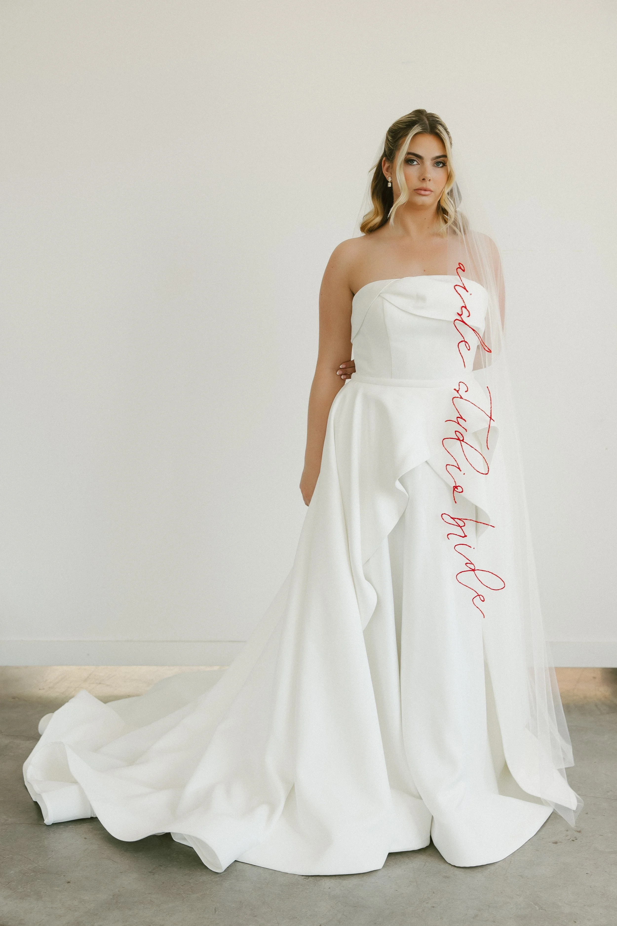 A woman stands in a white wedding dress. The dress is strapless with an asymmetrical neckline detail and delicate ruffle detail on the skirt.