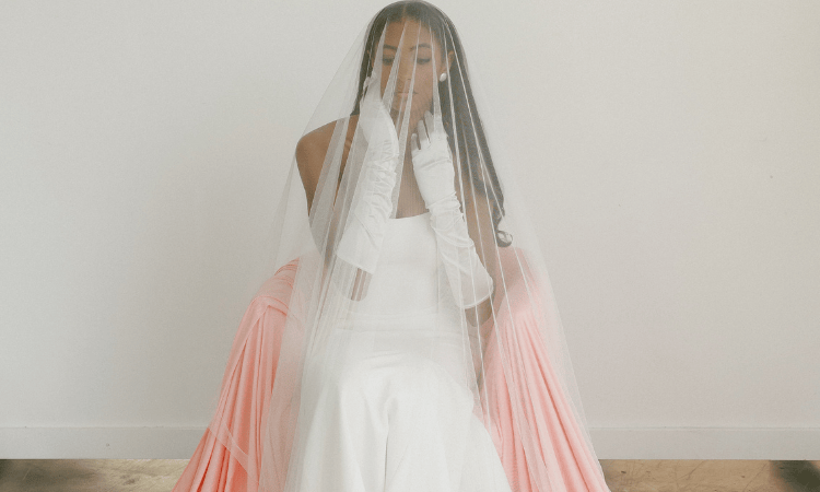 Bride in a white gown sits gracefully, draped in a long veil and wearing elegant gloves. The soft pink sash adds a touch of color and serenity.