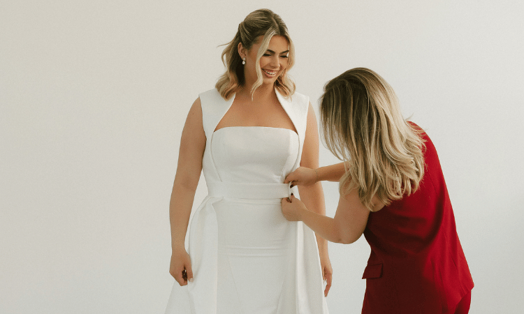 Bridal stylist adjusting the waist of a fitted wedding dress while the bride smiles during a bridal boutique fitting